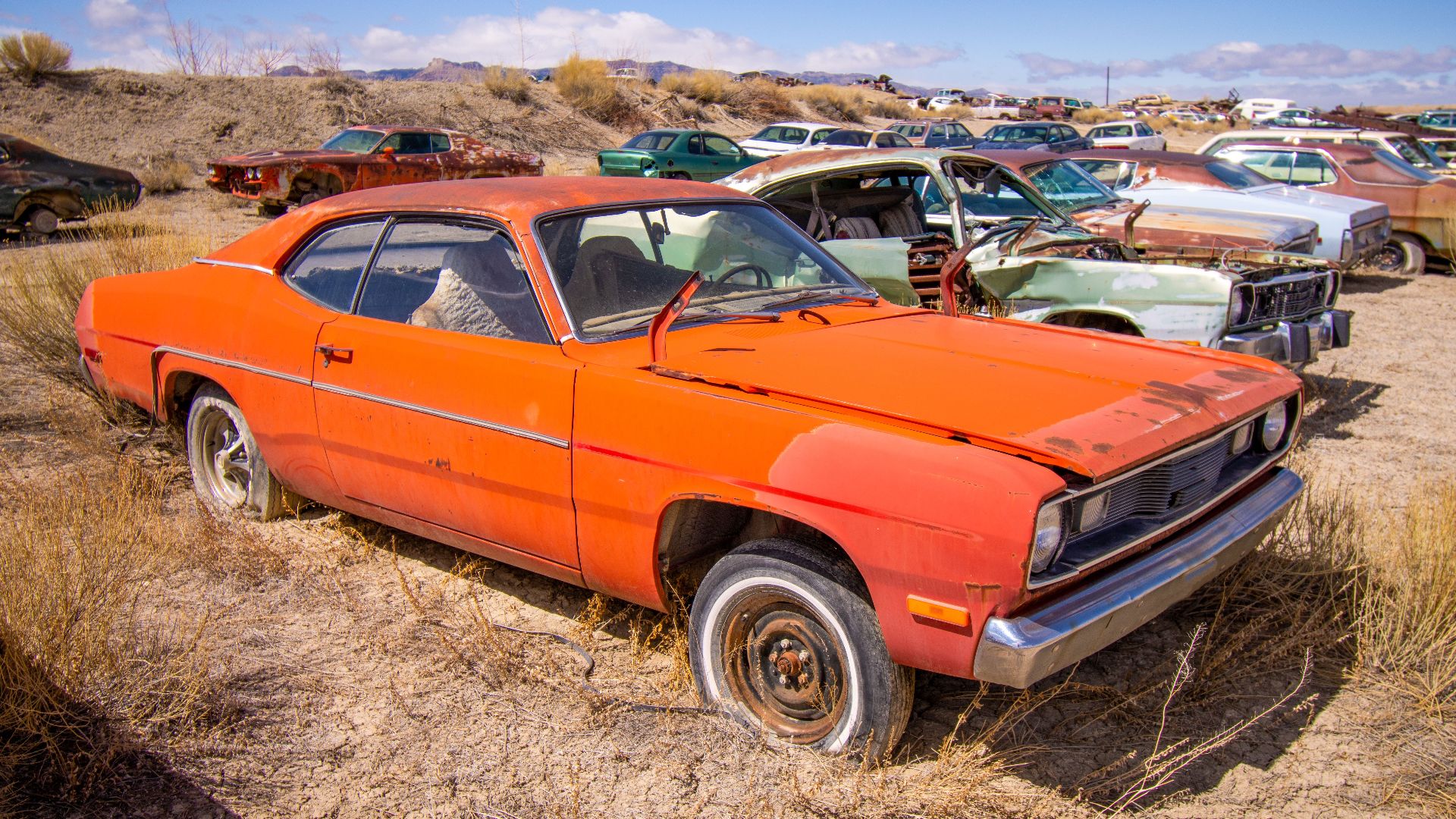 An old, rusted car sits abandoned in a scrapyard under the sunny desert sky.