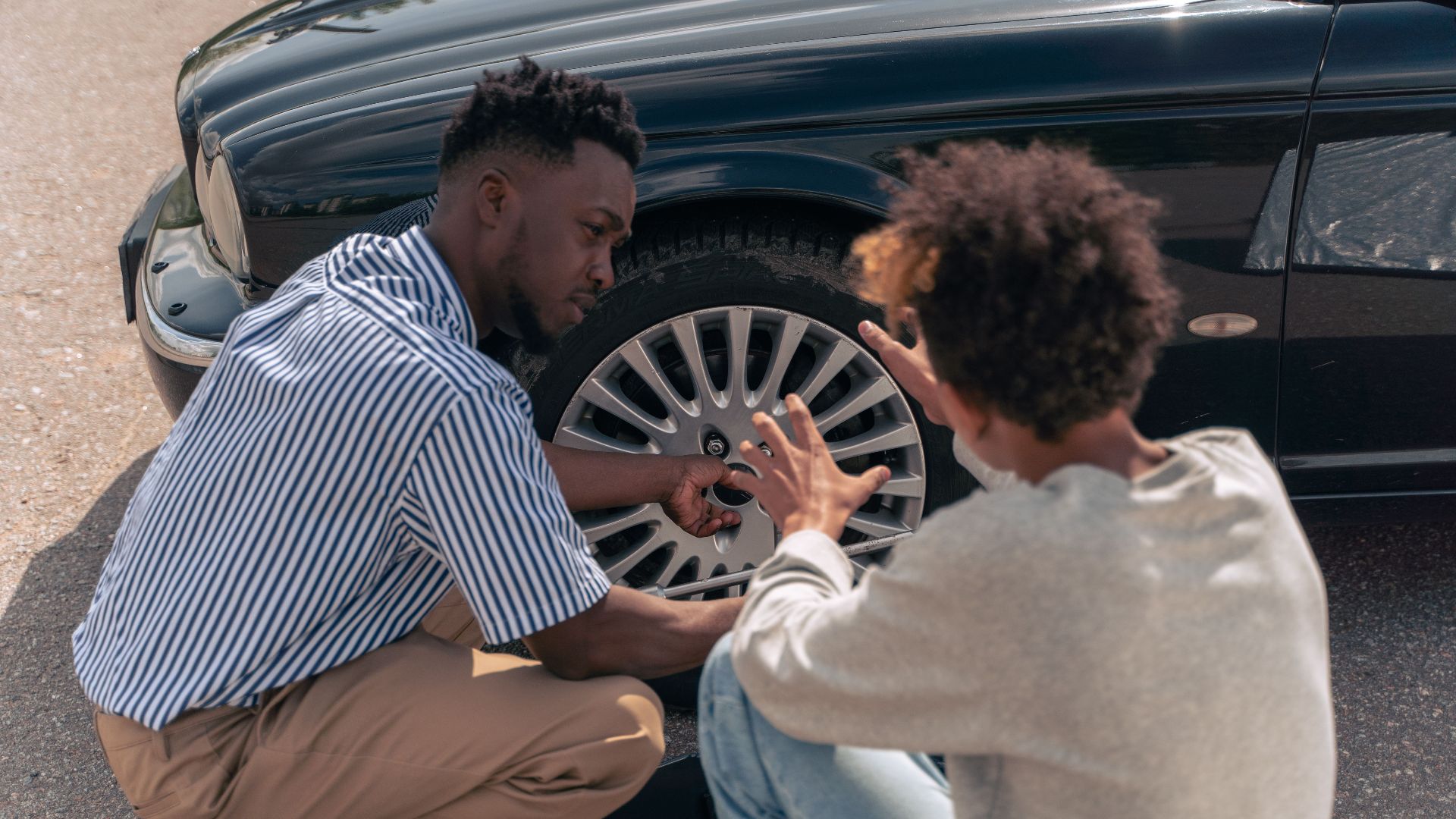 A father instructs his teenage son on how to change a car tire outdoors.