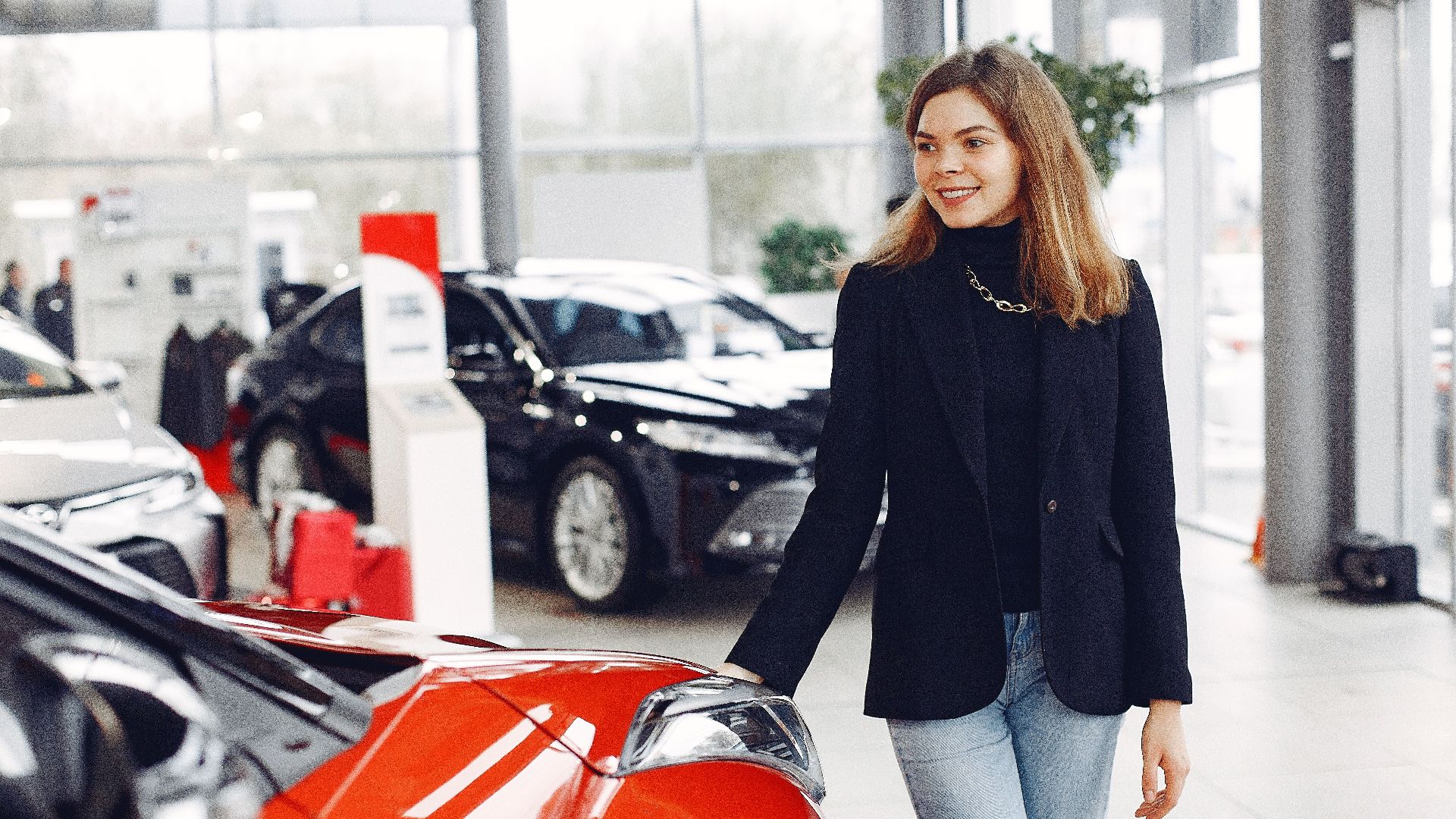 Happy stylish young female in jeans and black jacket standing near modern red shiny car in contemporary car showroom and looking at purchase dreamily