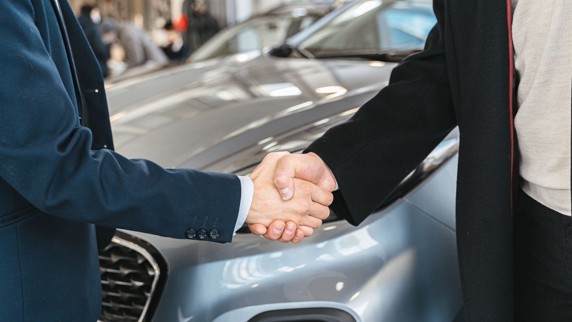 A close-up of two people shaking hands in front of a car in an indoor setting.