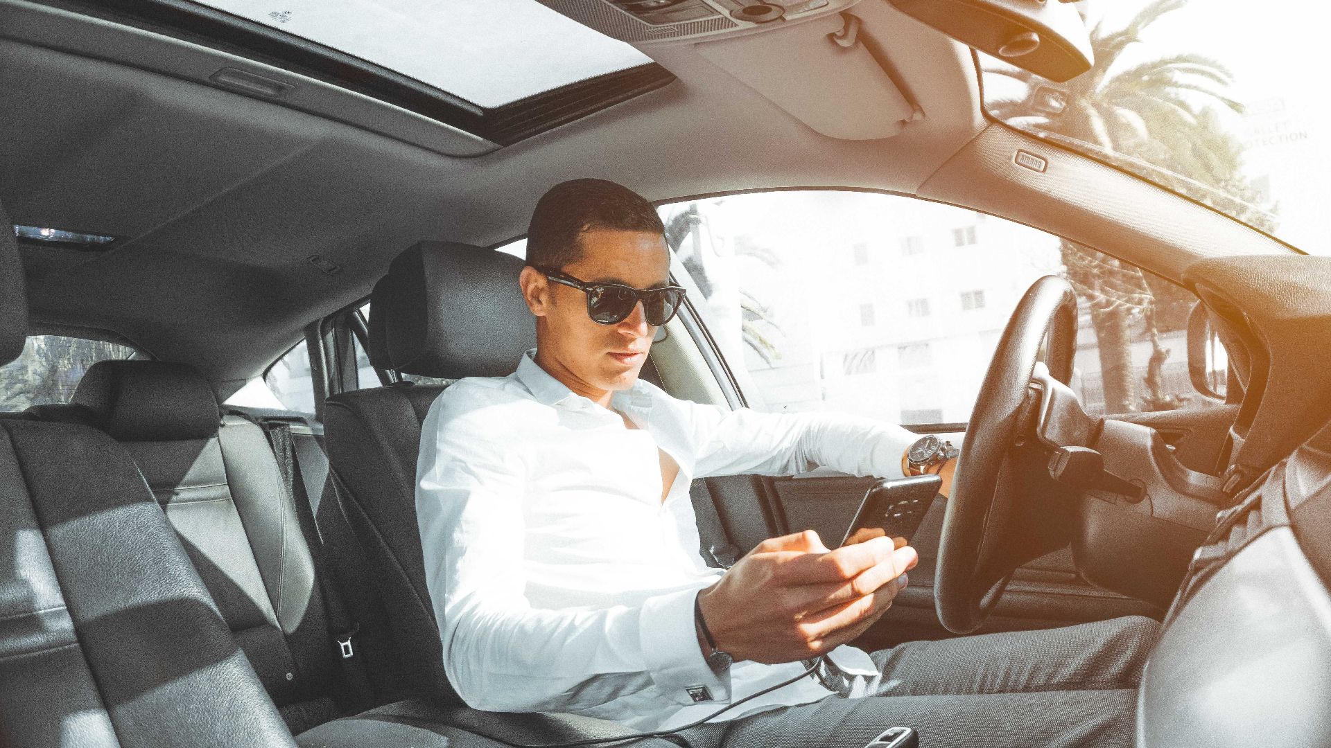 A man wearing sunglasses texts on his phone while driving a car in Morocco.