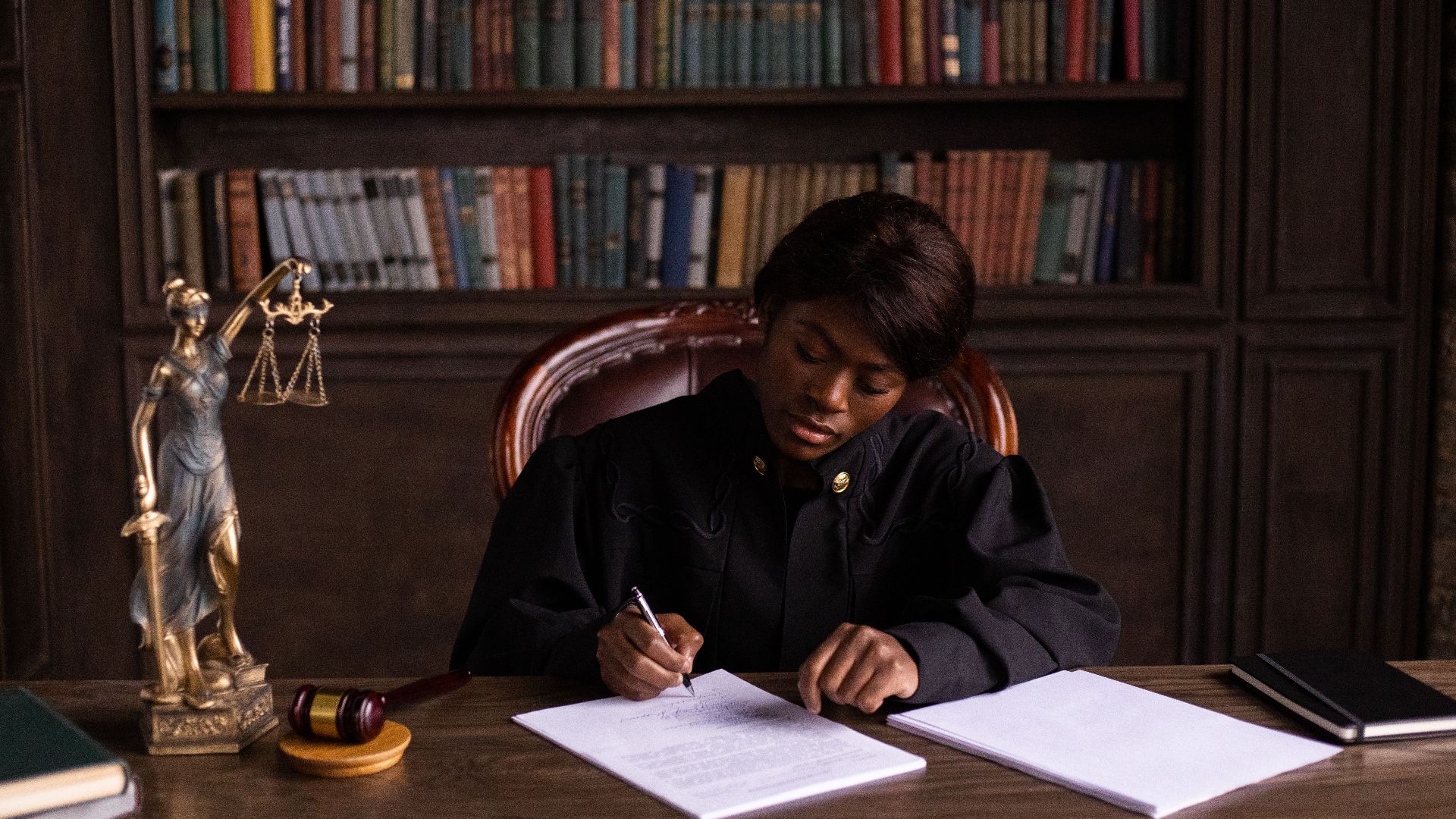 A judge in robes writing on a document at a desk in an office library with law books.