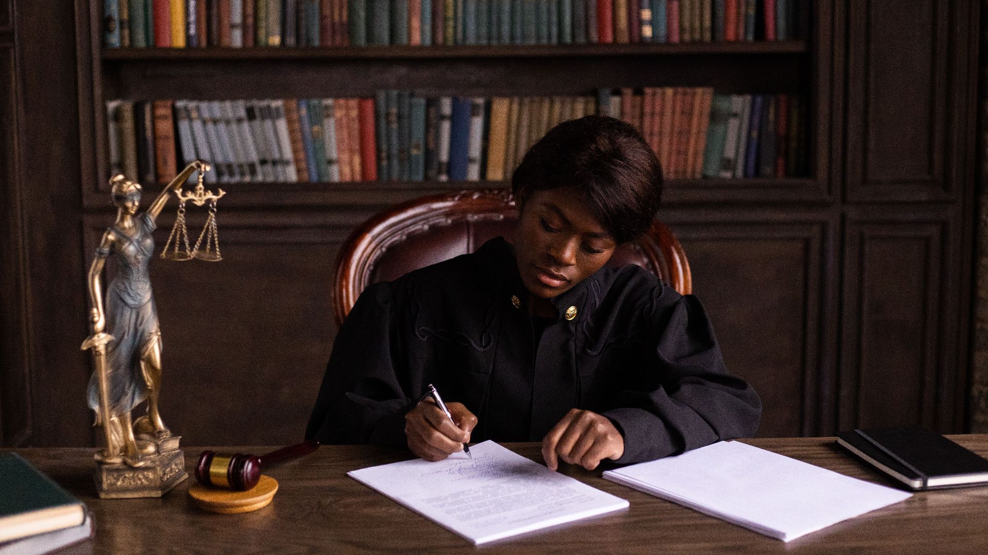 A judge in robes writing on a document at a desk in an office library with law books.