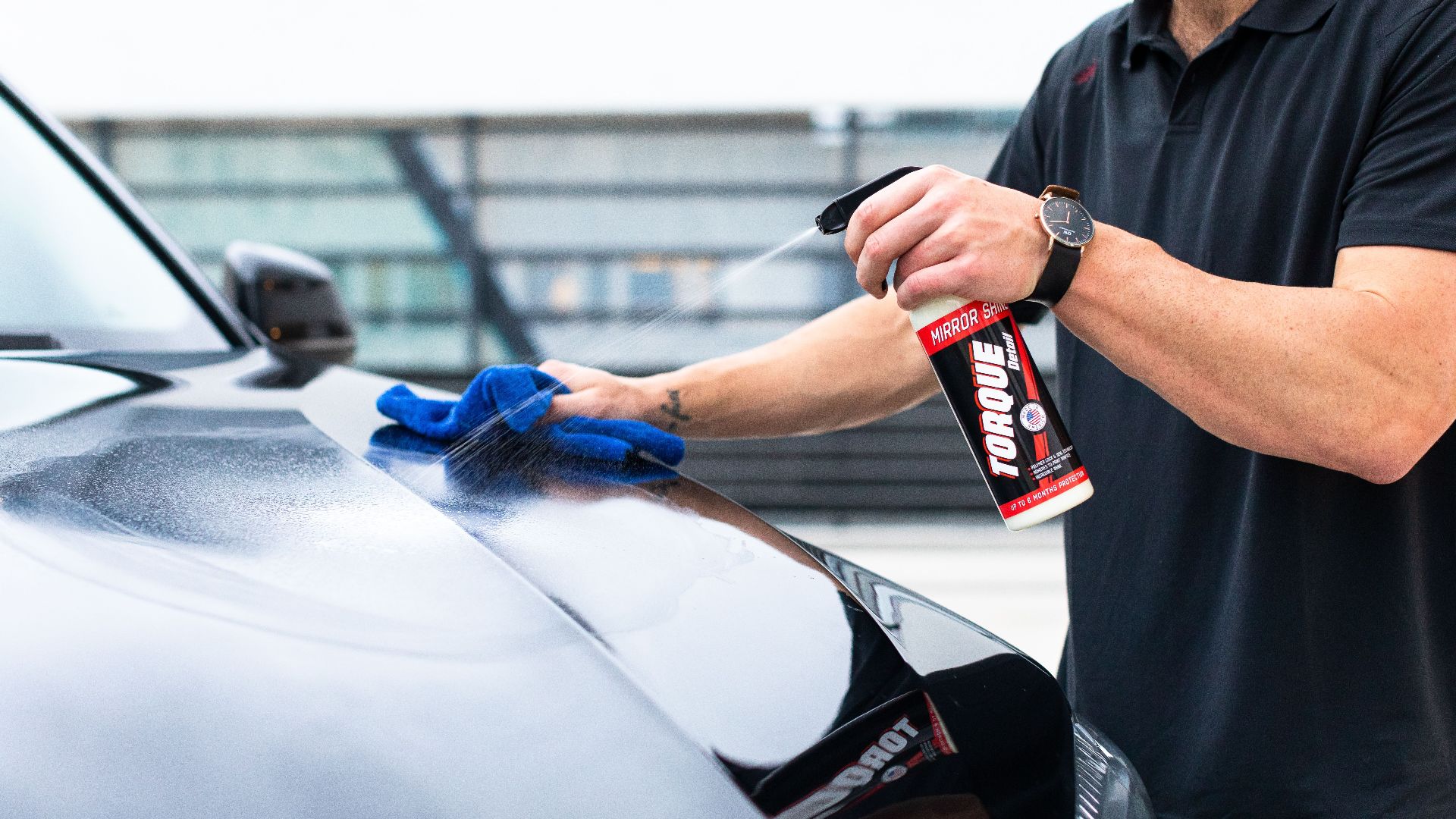 Close-up of a man cleaning a car hood with spray and microfiber cloth outdoors.