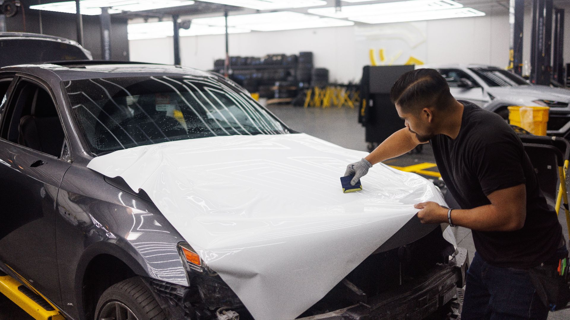 Man applying vinyl wrap on car hood in an automotive workshop setting.