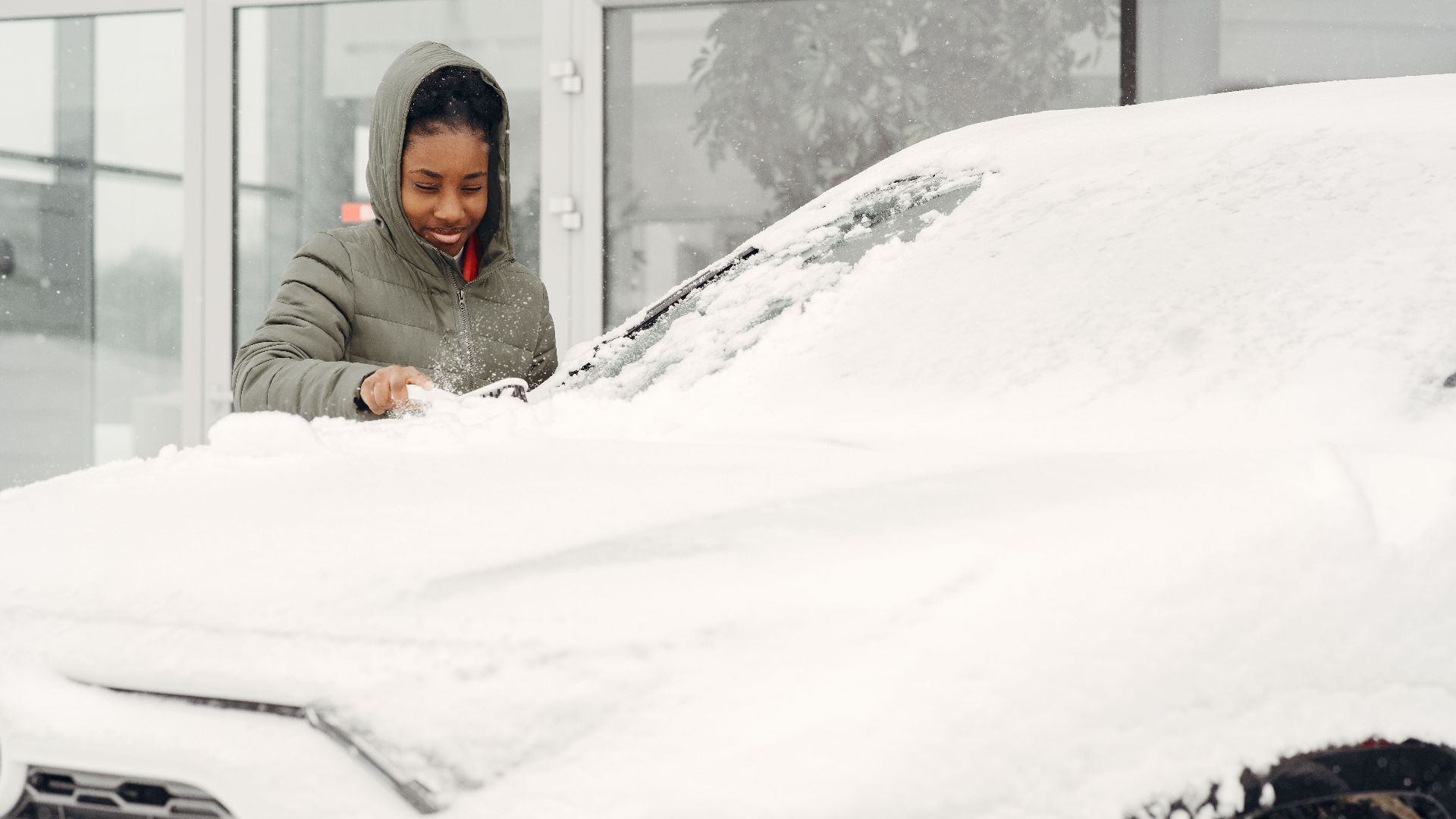 A woman in a gray coat clears snow from a car with a smile during a winter day outdoor.