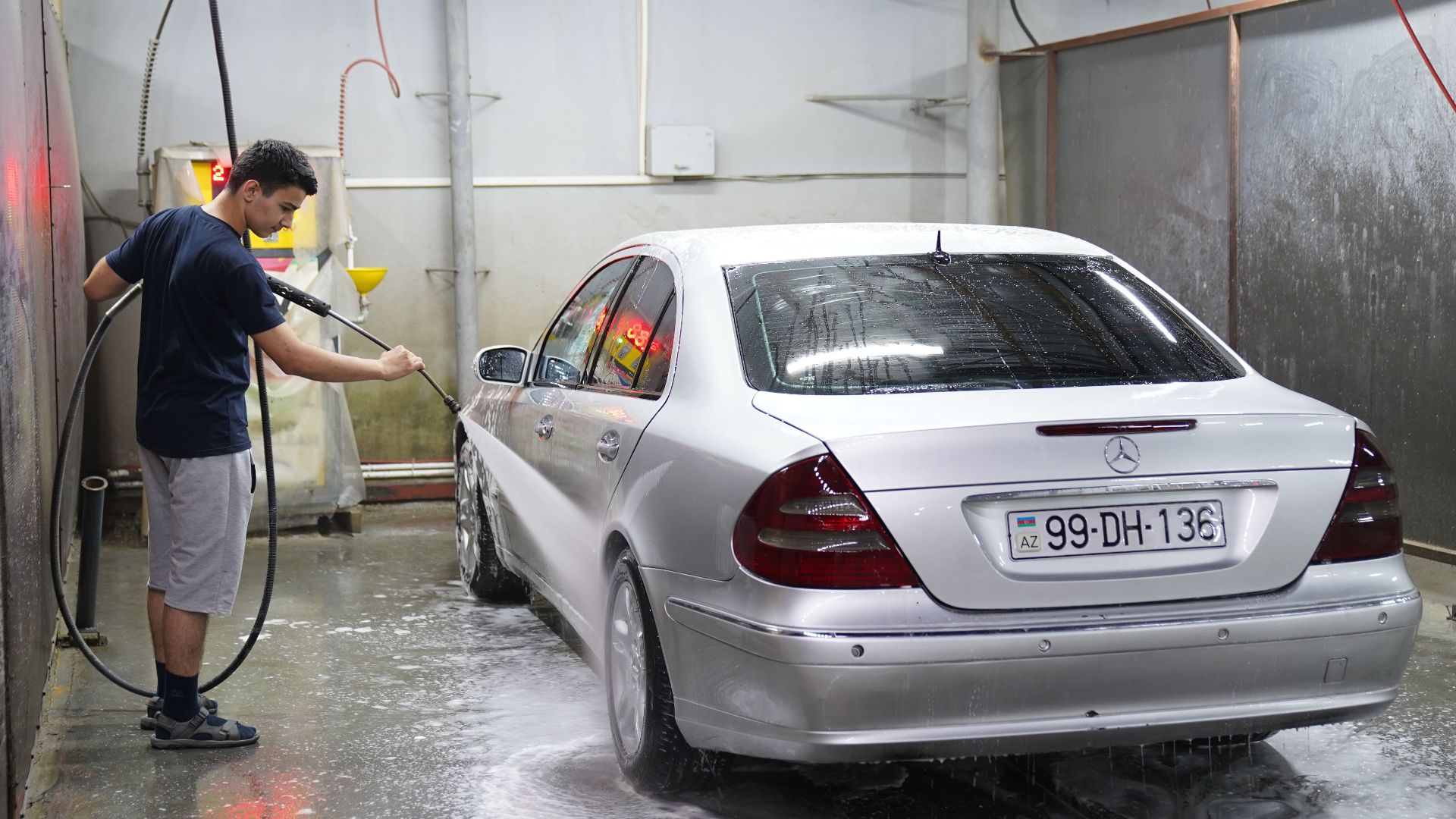 A young man using a hose to wash a silver car inside an indoor car wash facility.