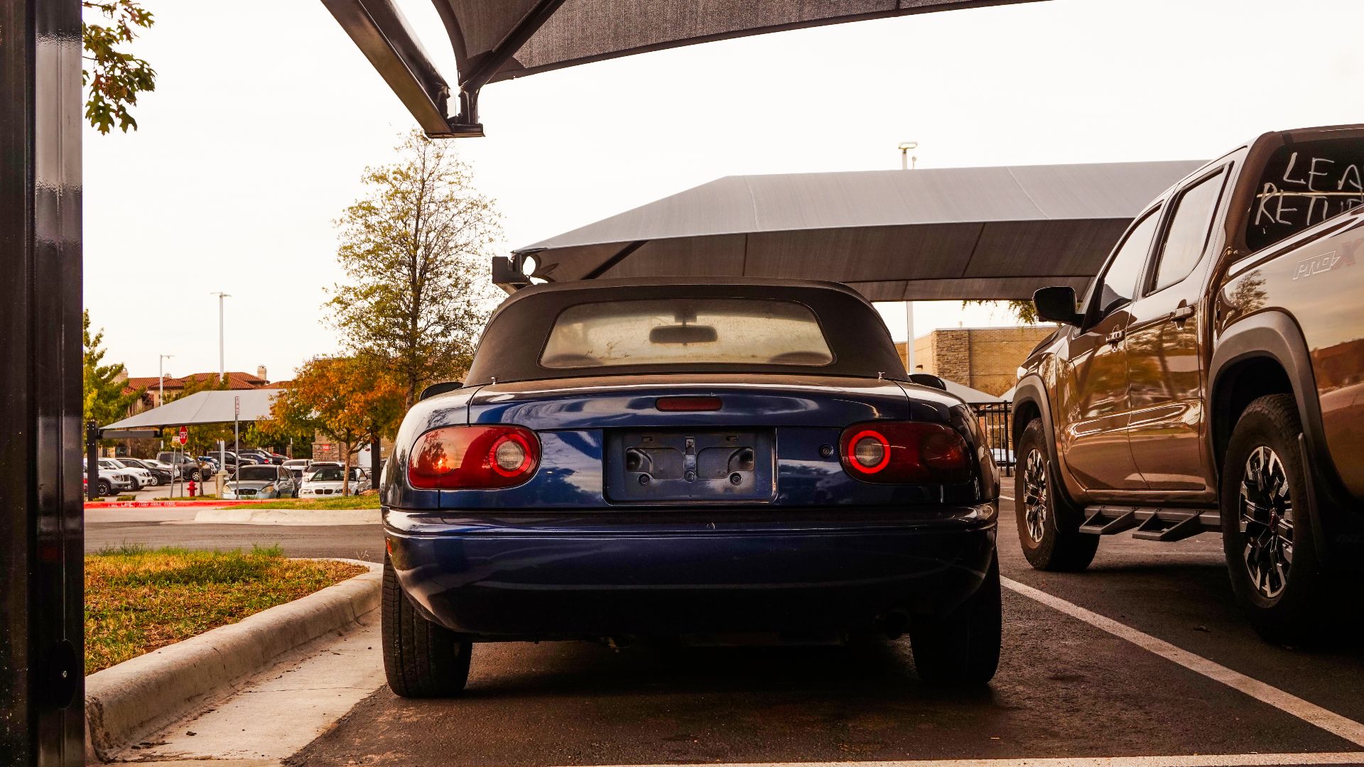 Rear view of a vintage blue convertible car parked beside a pickup in a city parking lot.