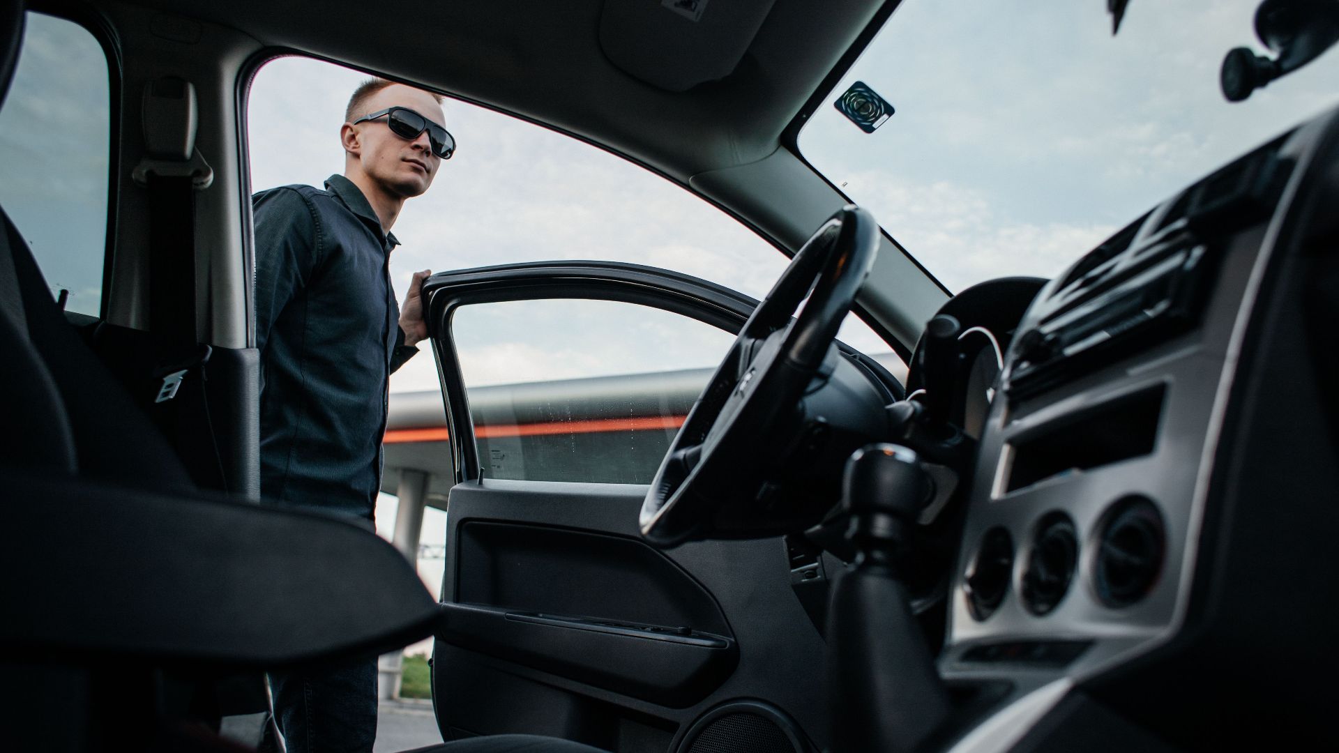 A man in sunglasses stands by an open car door, viewed from the interior.