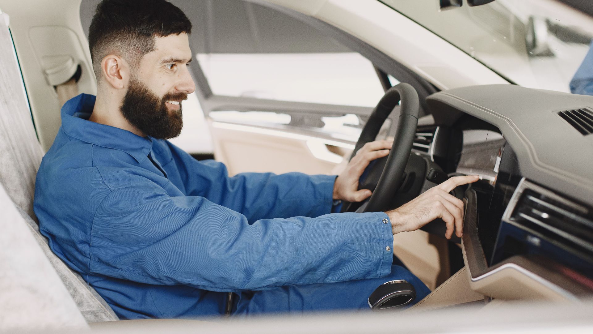 Mechanic in blue coveralls interacts with car dashboard, smiling and focused.