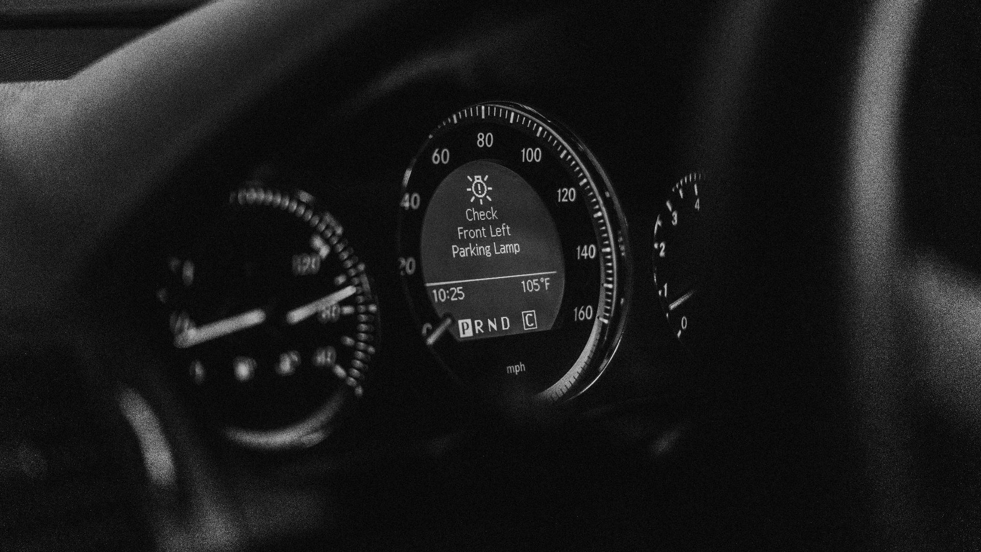 Close-up of a car dashboard in black and white with a parking lamp warning light.