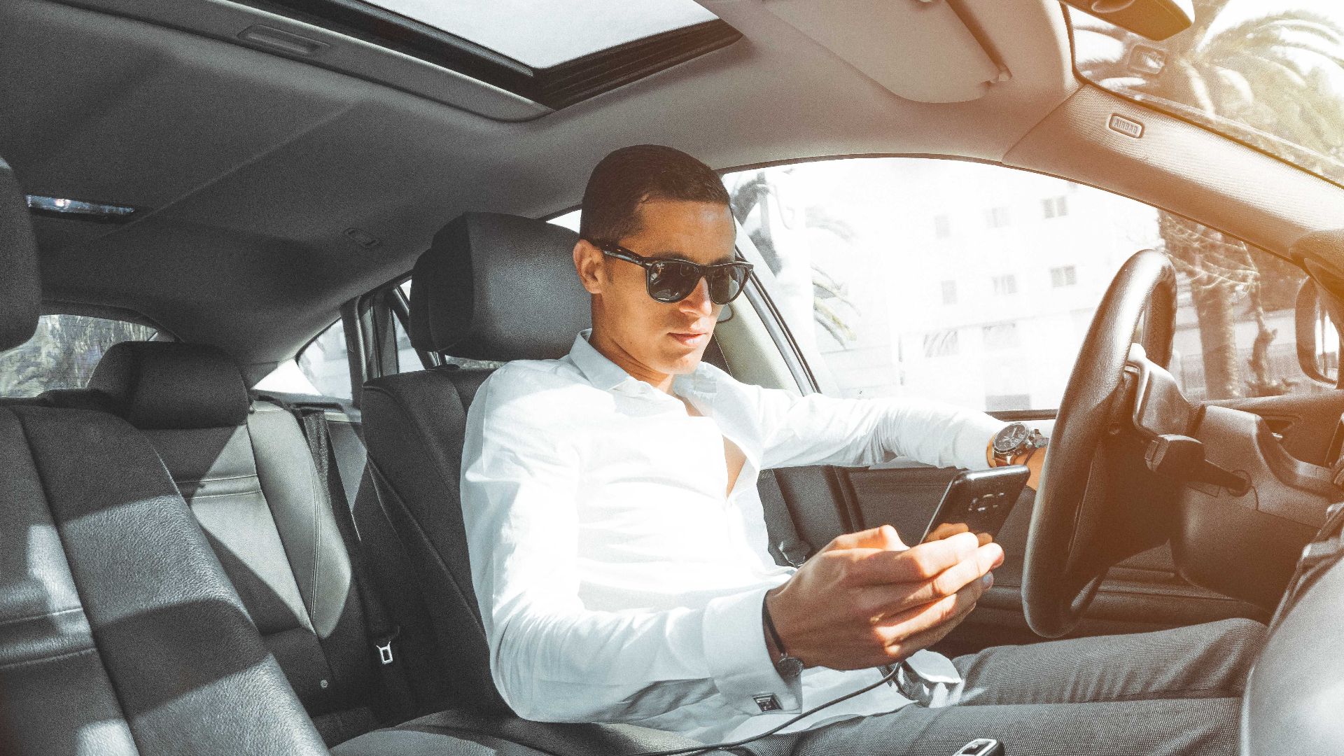 A man wearing sunglasses texts on his phone while driving a car in Morocco.