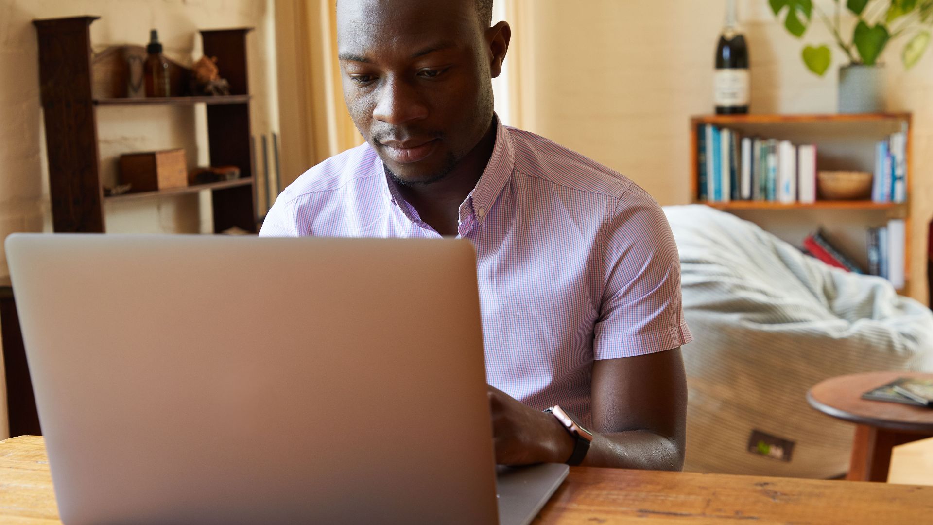 Concentrated African American male typing information on netbook at wooden table in cozy living room