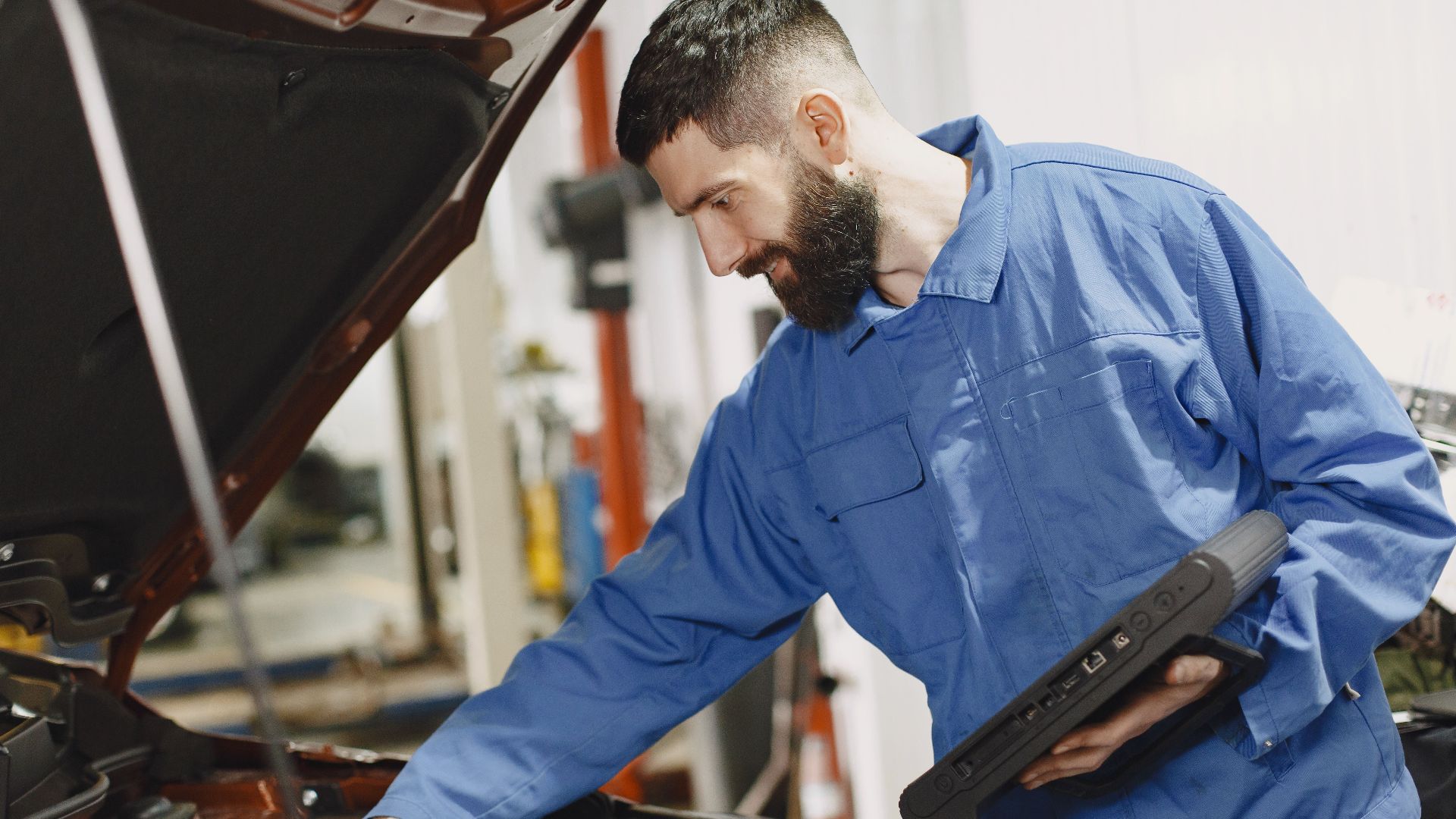 An auto mechanic in blue coveralls inspecting a car's engine in a garage.
