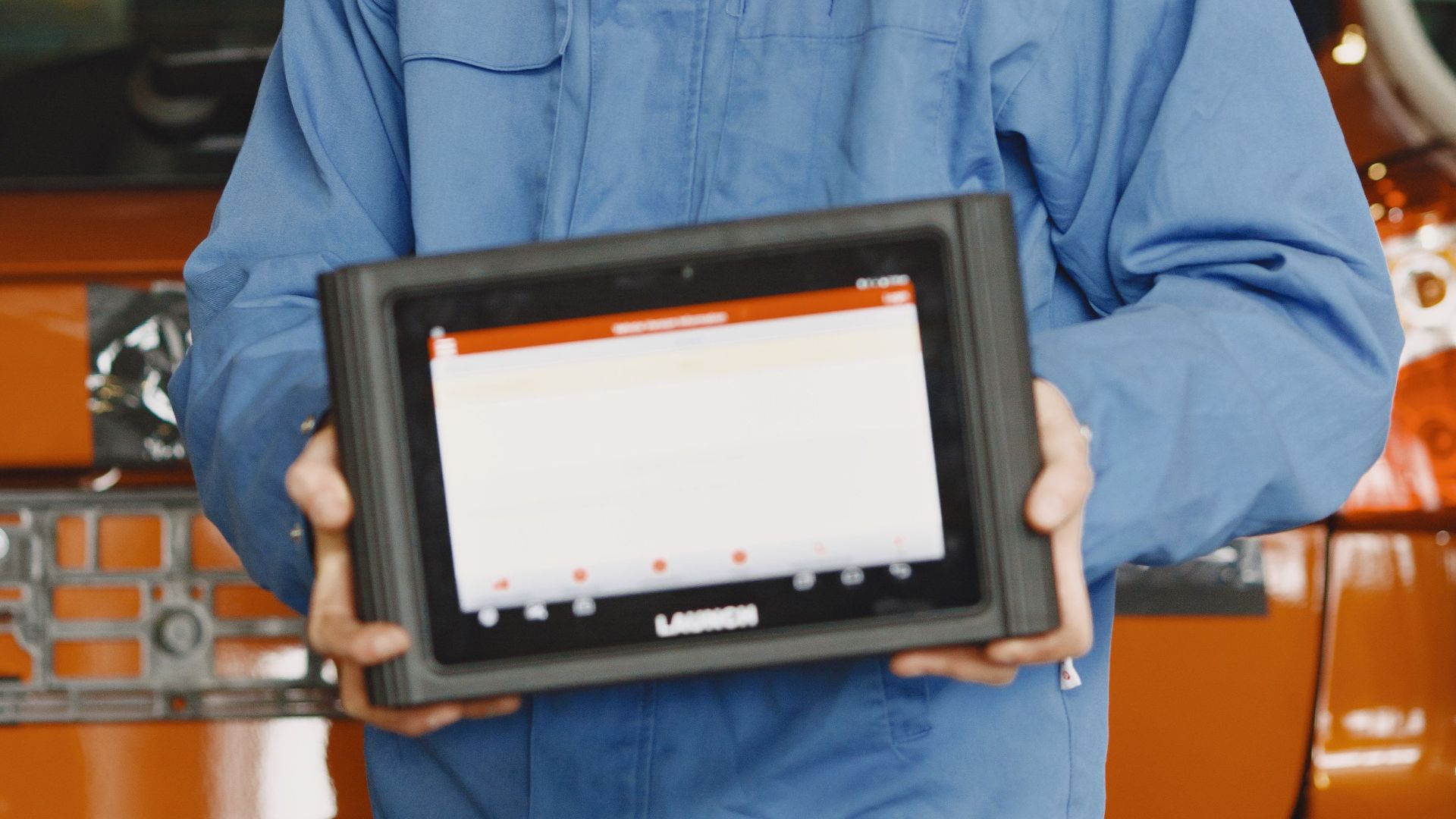 Auto mechanic in blue coveralls holding a diagnostic tablet in an auto repair shop.
