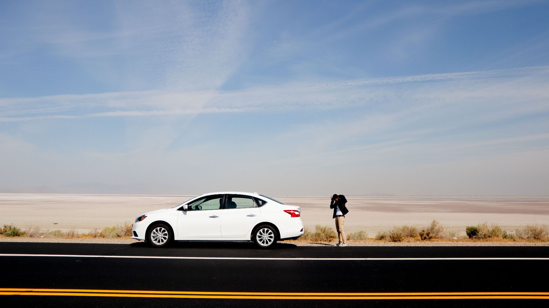 A white car parked on a desert road with a photographer capturing the vast landscape under a clear sky.