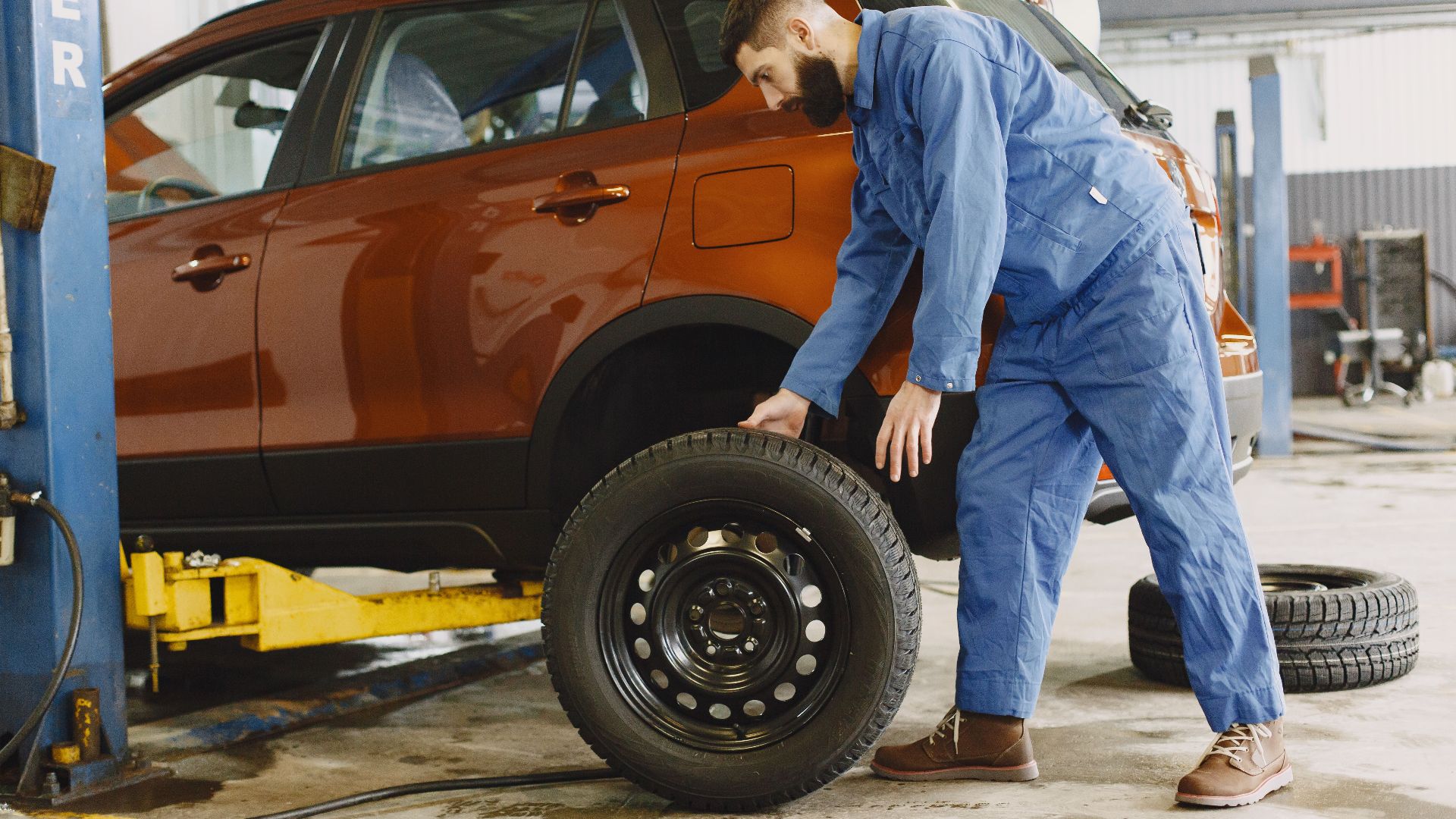 Mechanic in blue coveralls replacing tire on red SUV in repair shop garage.