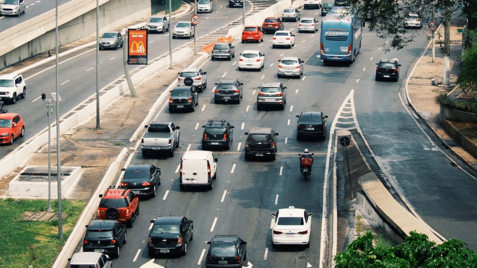 Cars and buses navigate a busy city road surrounded by tall buildings and lush greenery.
