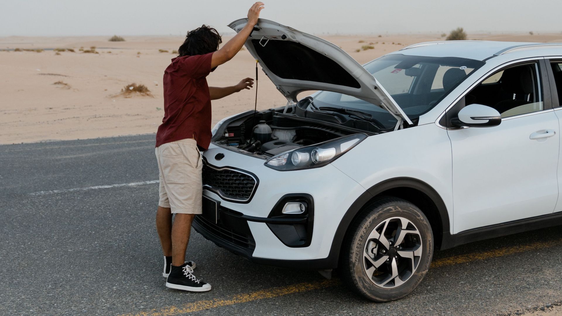 A man inspecting the engine of a car by the roadside in a desert setting.
