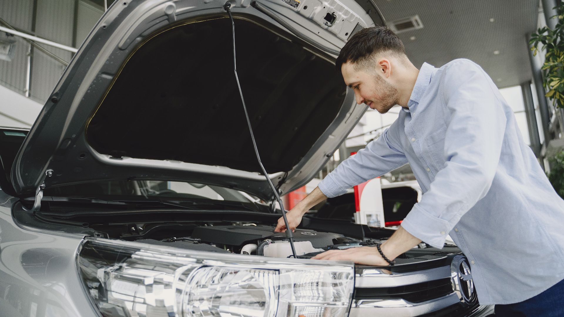 A man in a blue shirt examines a car's engine in a modern vehicle showroom.