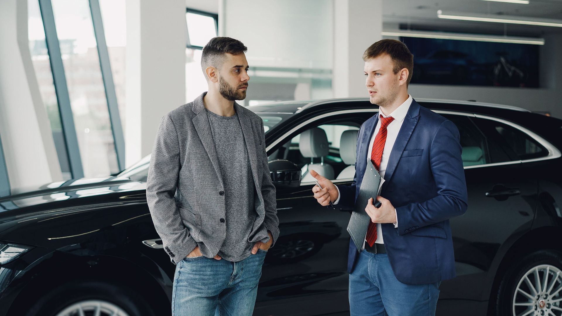 Two men discussing business in a car dealership, standing near a vehicle.