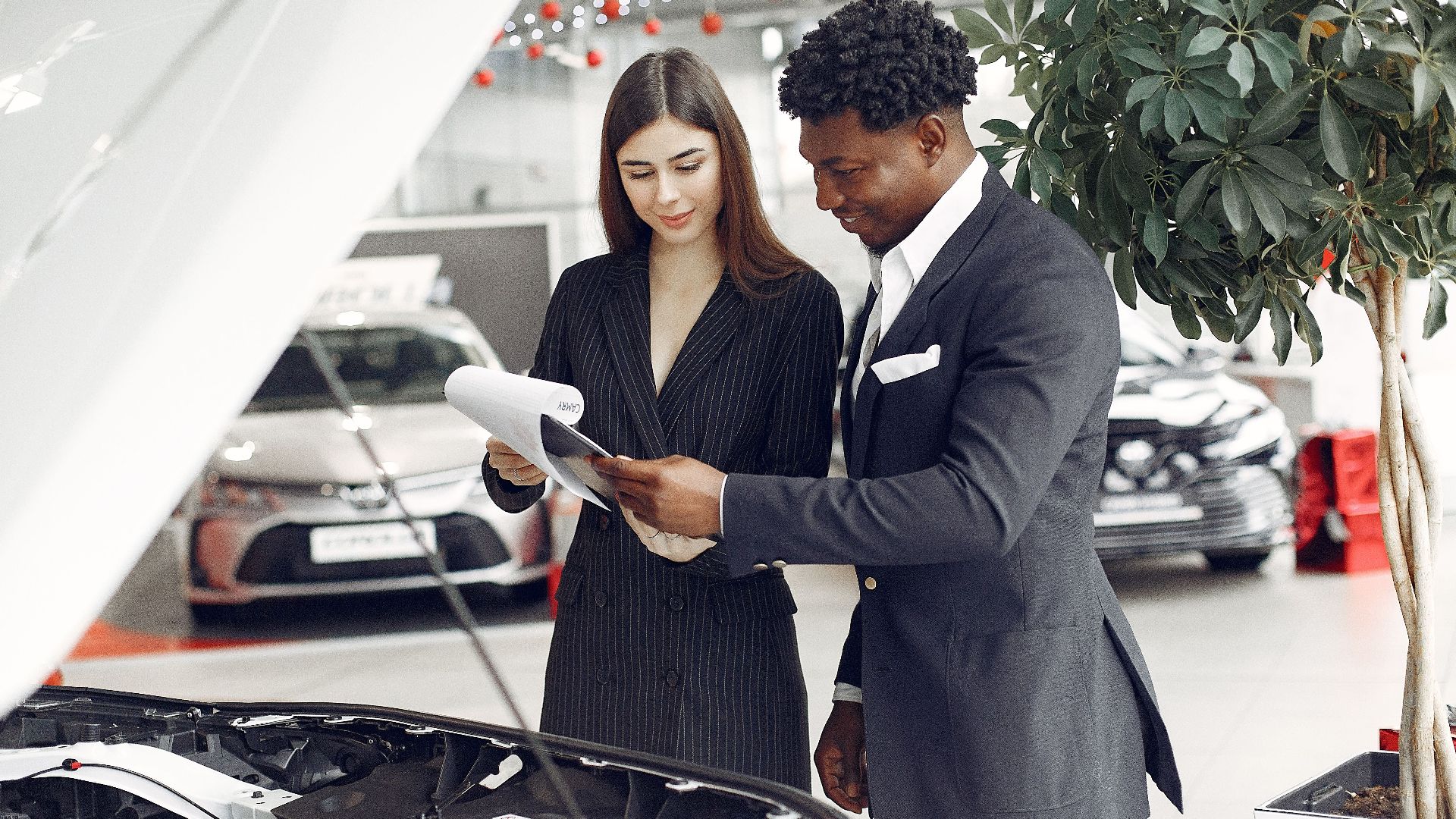Confident smiling successful black businessman in classy formal suit standing near open motor hood in car showroom and discussing car characteristics with gorgeous female dealer