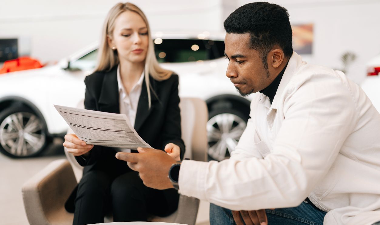 Medium shot of female manager consulting doubtful black buyer, giving purchase agreement for review at showroom. Thoughtful African client receiving car purchase contract before signing in dealership