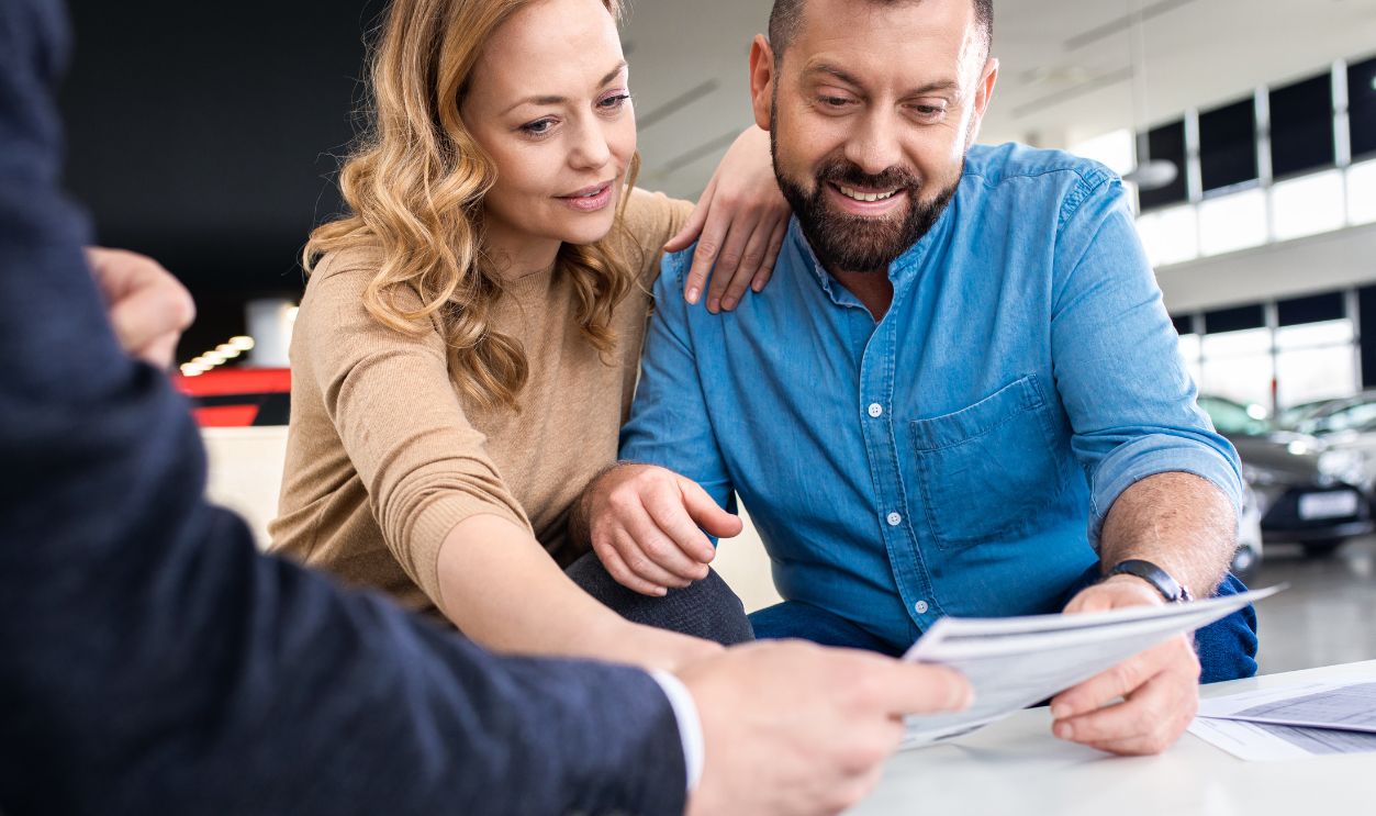 Mid-adult couple talking with sales agent and signing contract for new car in modern car showroom.