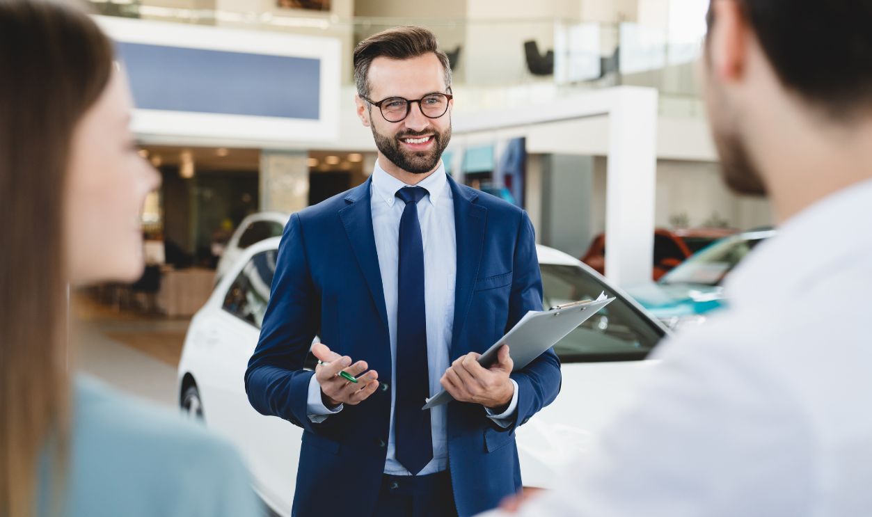 Focused photo of young male shop assistant explaining talking offering buying new car to young caucasian family husband and wife at auto dealer shop.