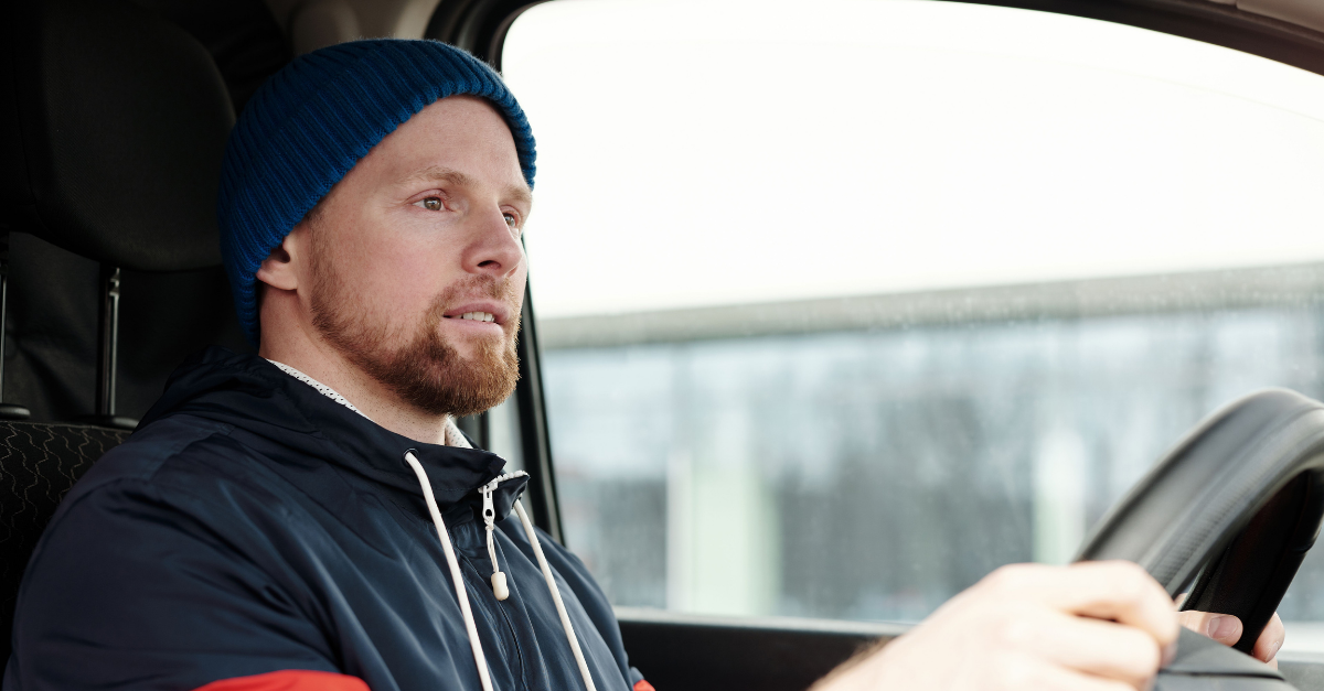 A bearded man wearing a hoodie and knitted cap driving a car, focused and attentive.