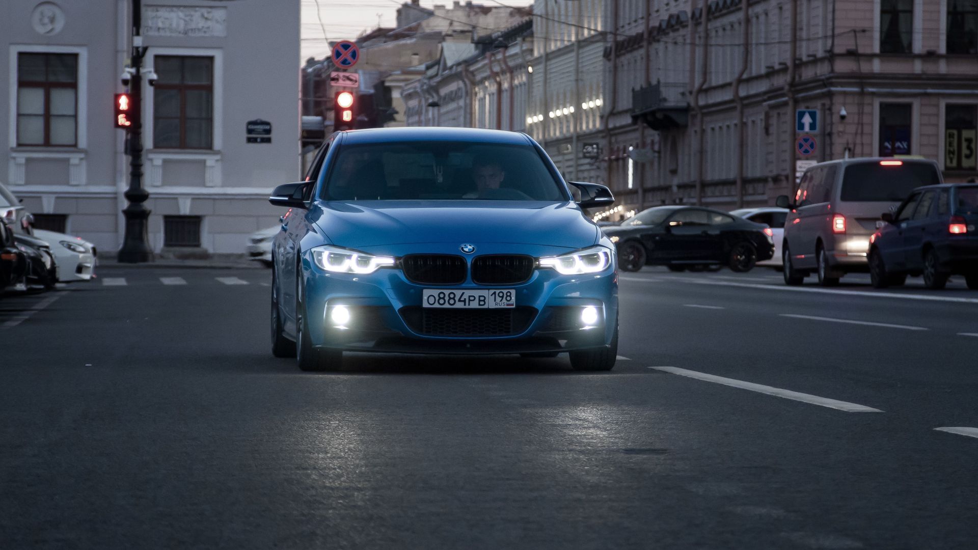 A blue car drives on a city street at dusk, showcasing urban life and motion.