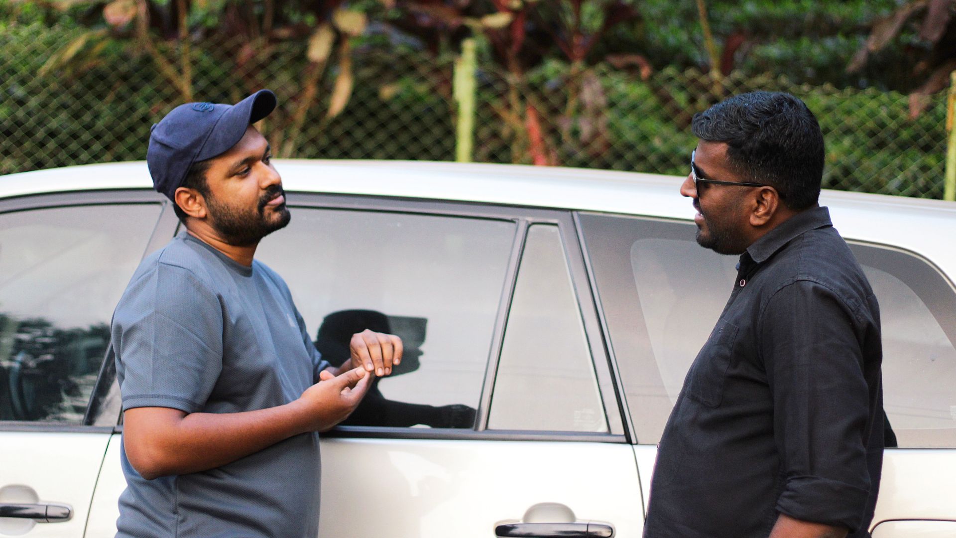 Two men having a casual discussion beside a car in an outdoor setting.