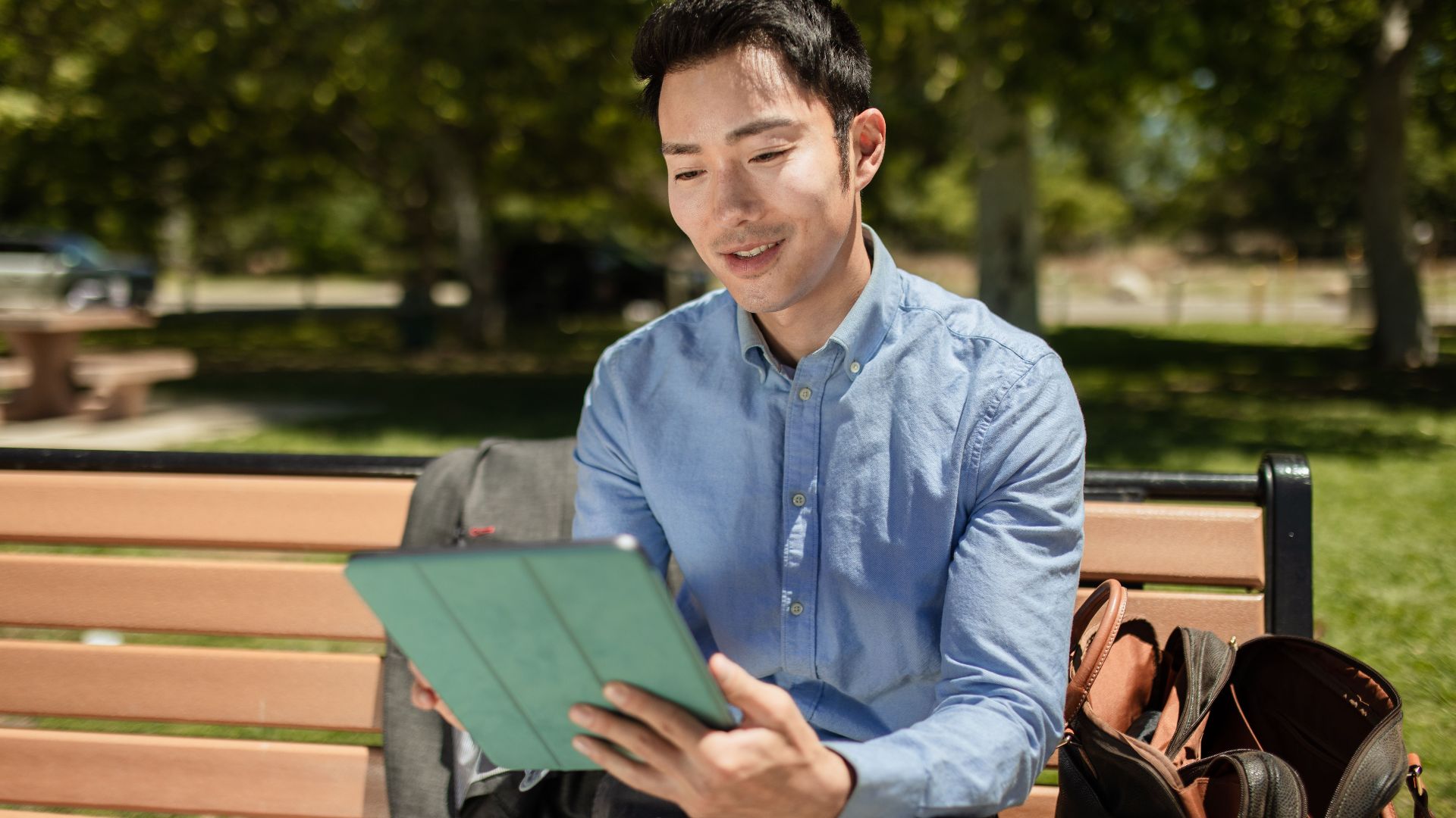 Asian man in corporate attire using a tablet on a park bench outdoors.