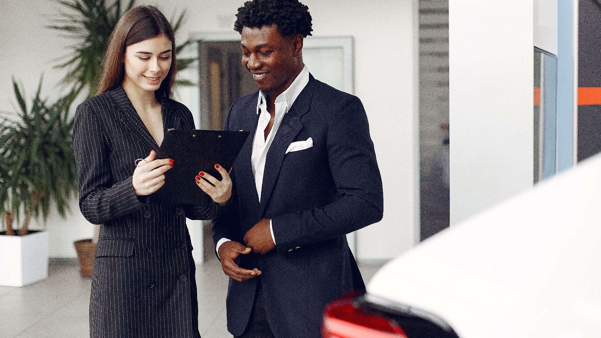 Cheerful multiethnic stylish female agent and smiling customer in formal suit discussing contract details while standing in modern car showroom in daylight