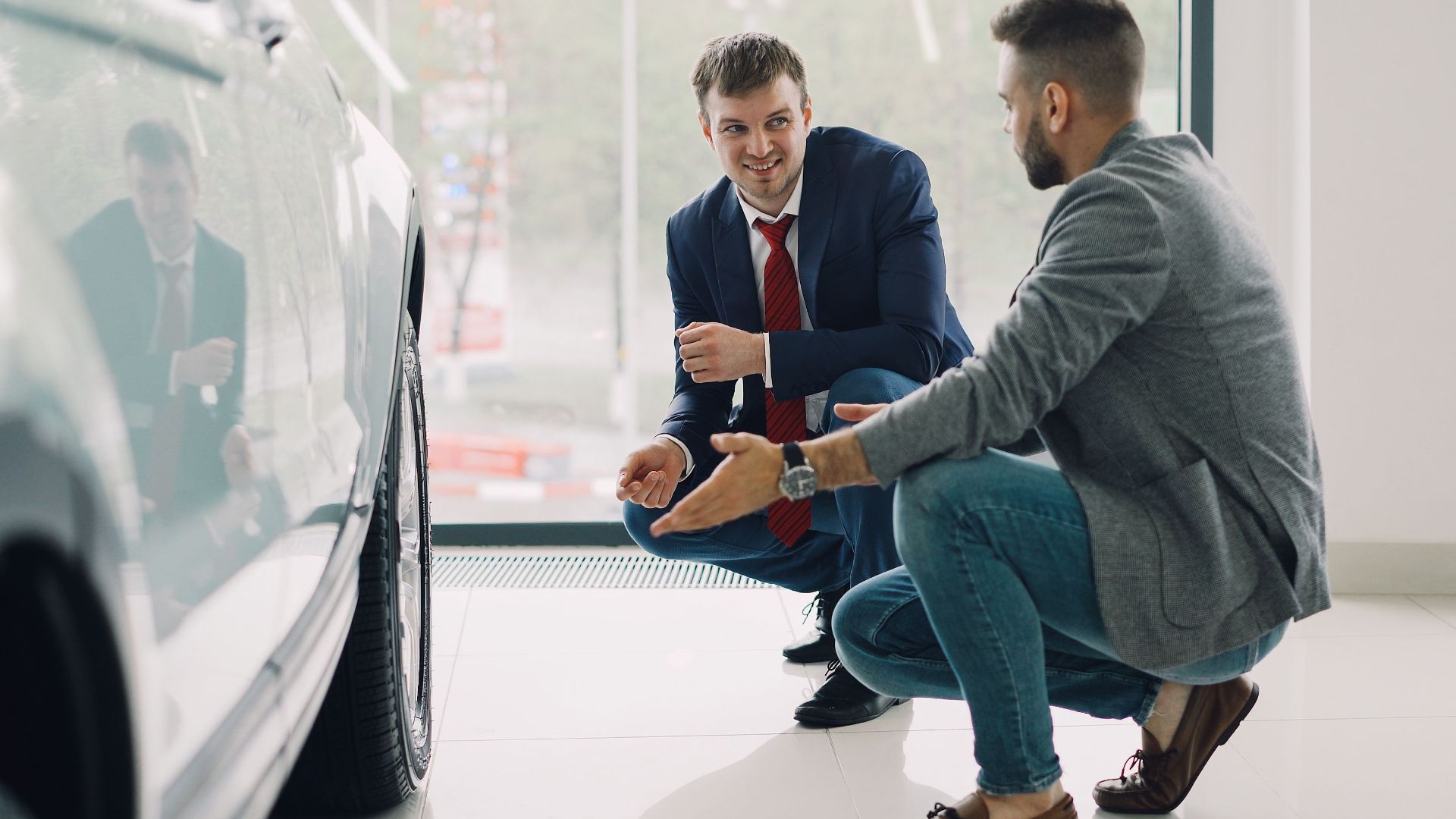 Two men discussing car features in a showroom, kneeling near a vehicle.
