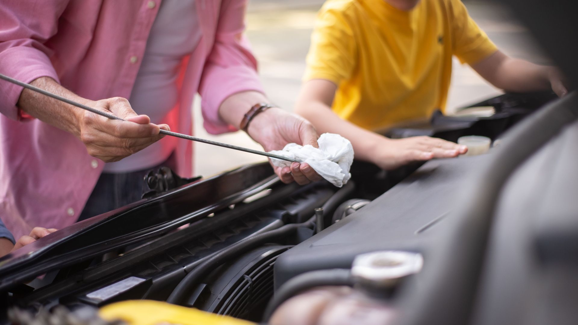 Father and son working on car maintenance, checking engine oil outdoors.