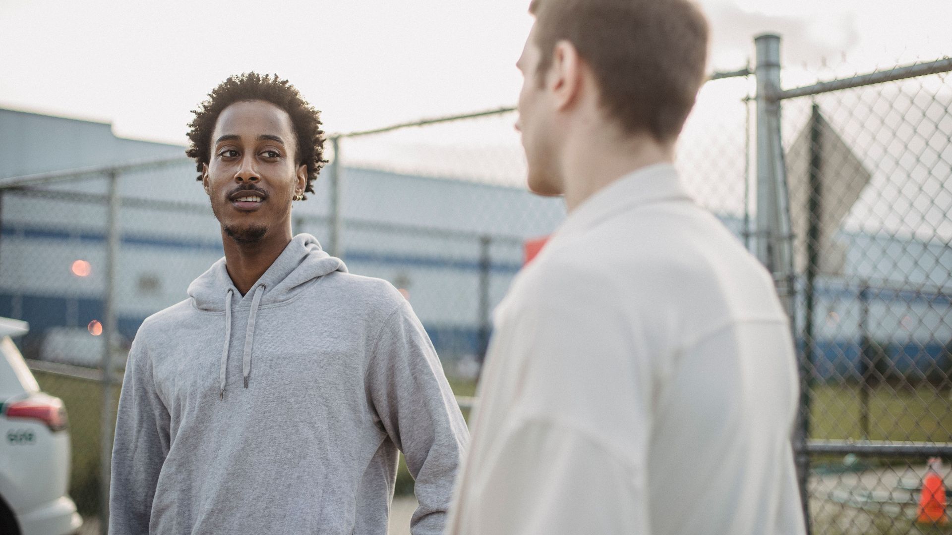 African American male standing with friend against metal grid near car on parking lot