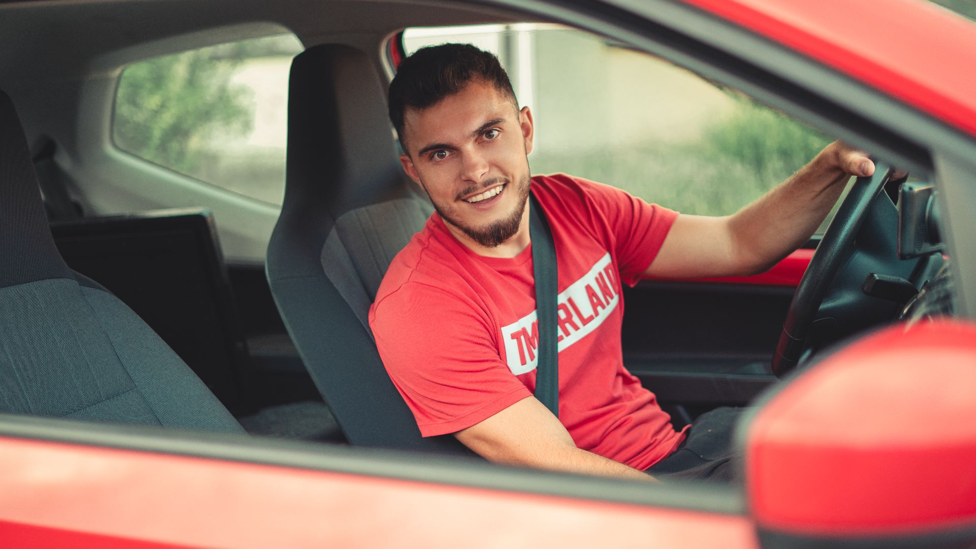 Smiling man driving a red car while wearing a seatbelt, showcasing safety and joy.