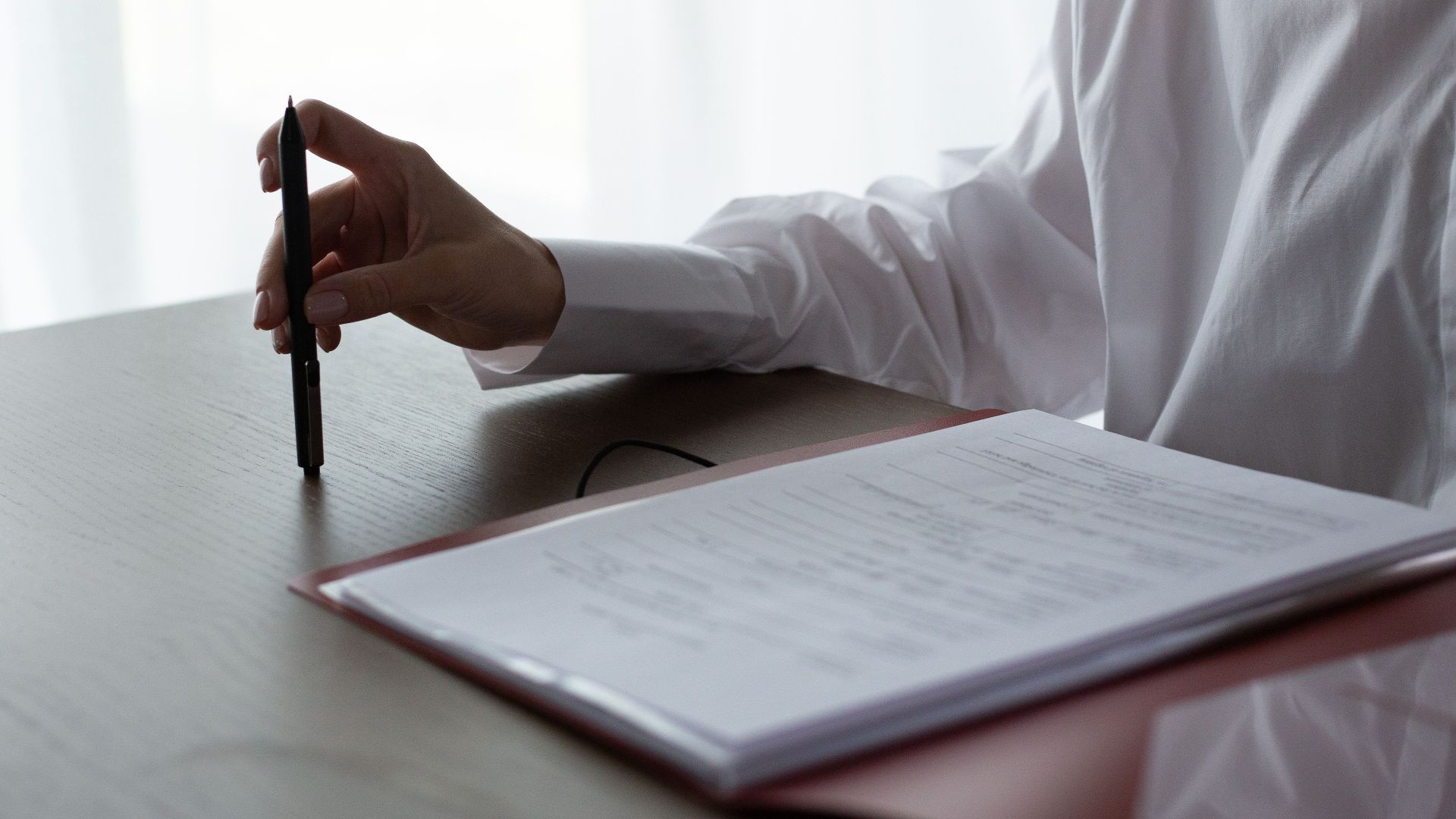 A woman examines documents, holding a pen over a table in a bright indoor setting.
