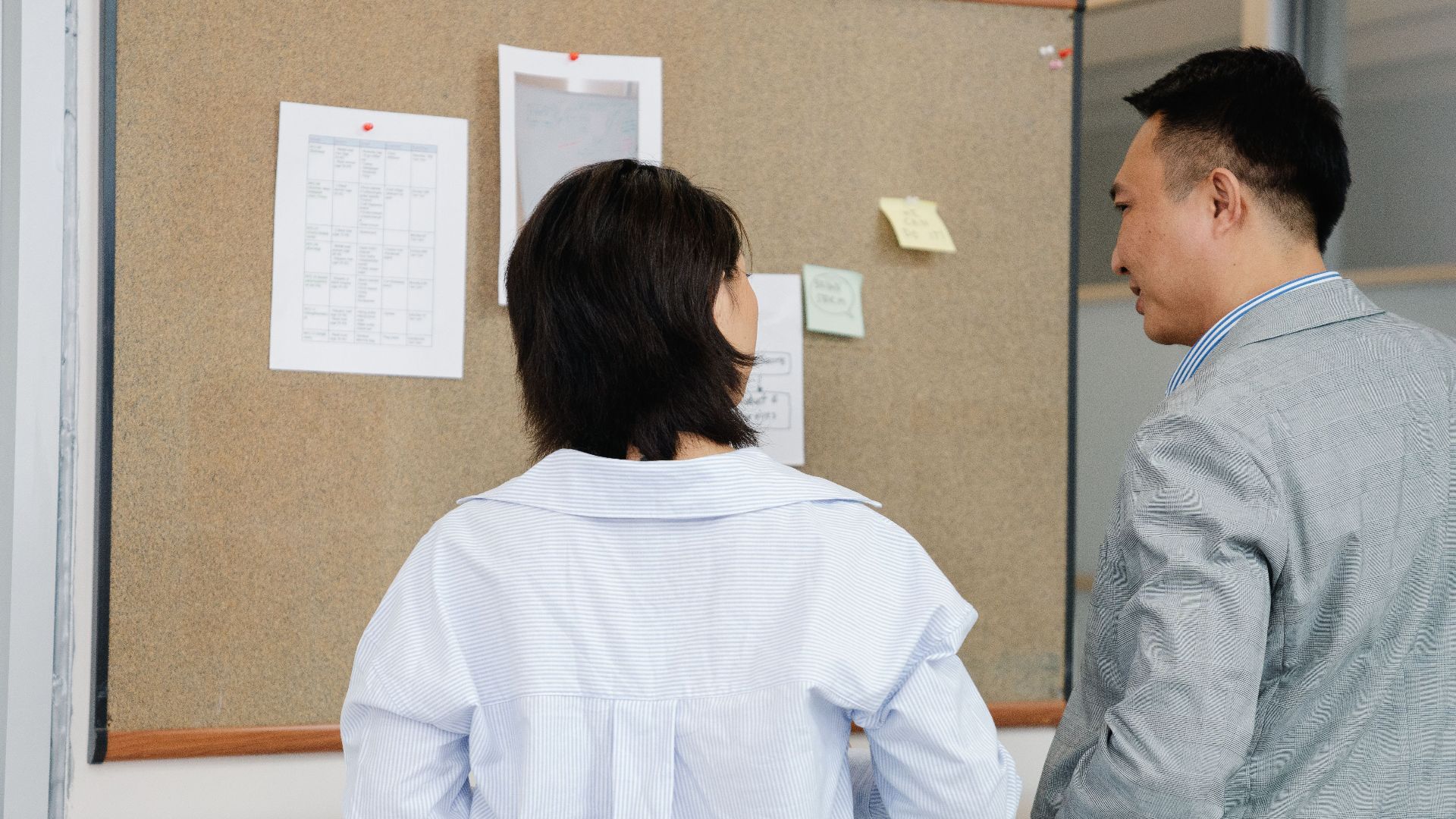 Two colleagues collaborate and discuss information on a bulletin board in an office setting.