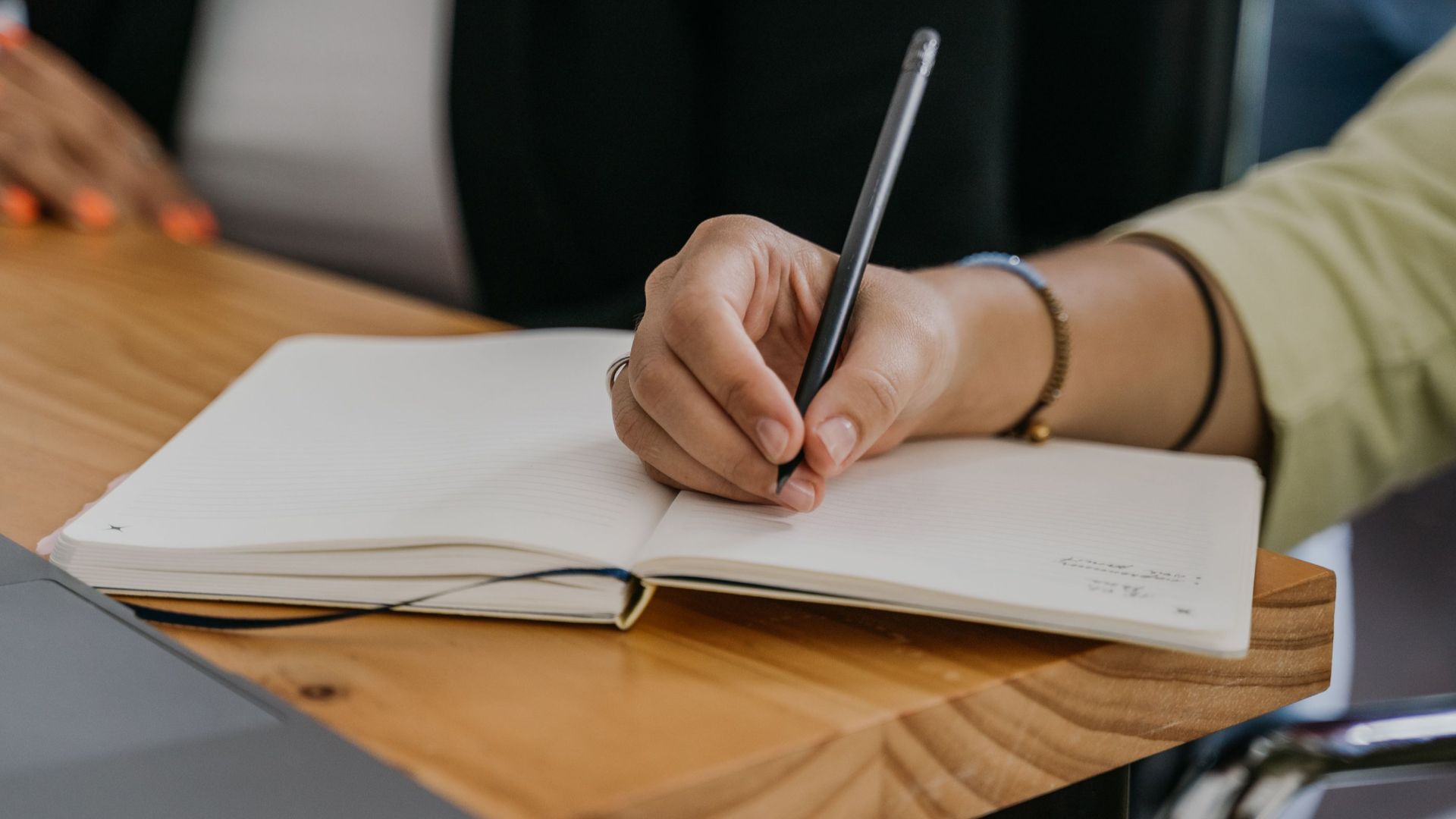 Close-up of hands writing in a notebook on a wooden table next to a laptop.