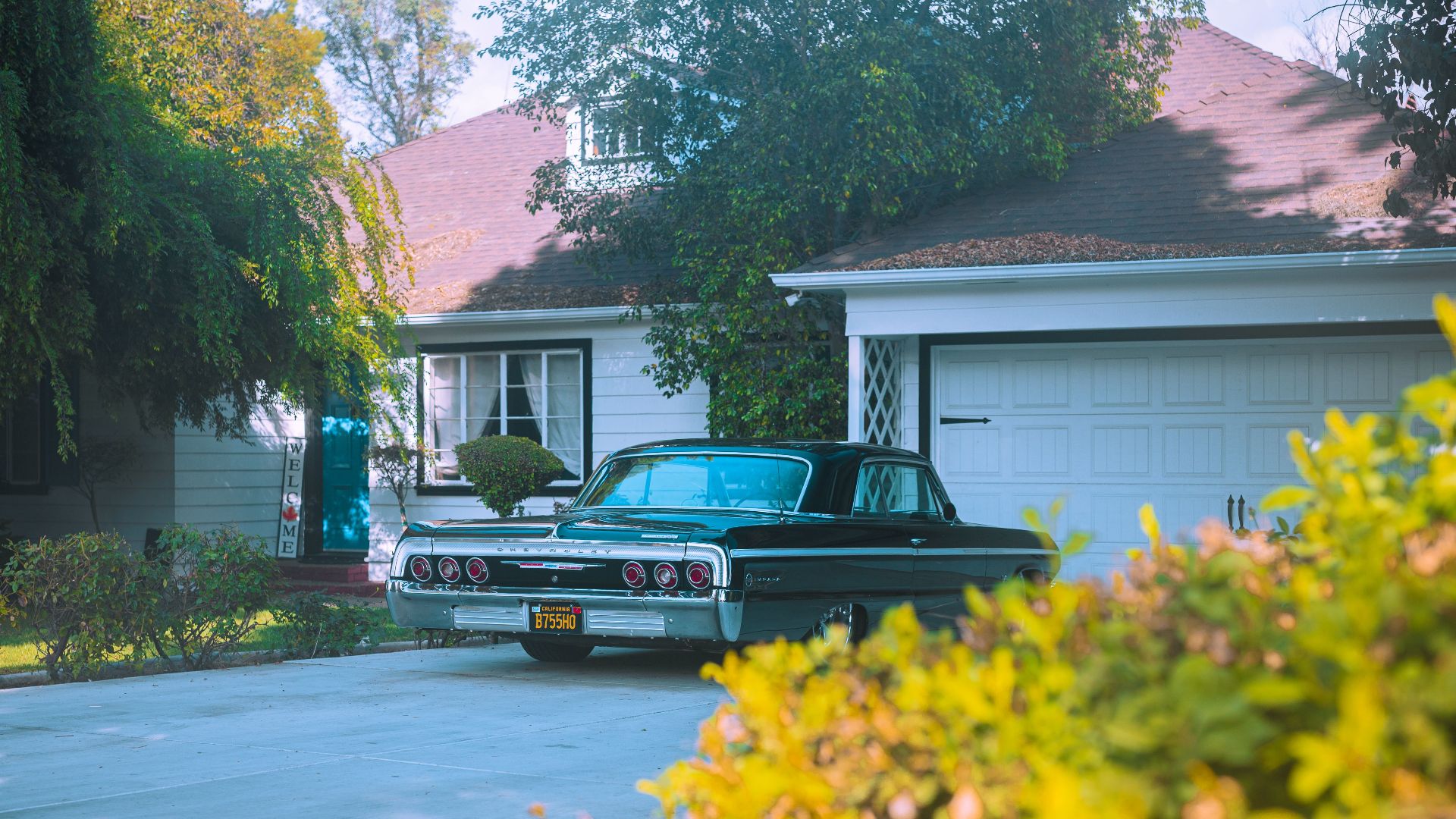 A classic black Chevrolet Impala parked in a suburban driveway surrounded by greenery.