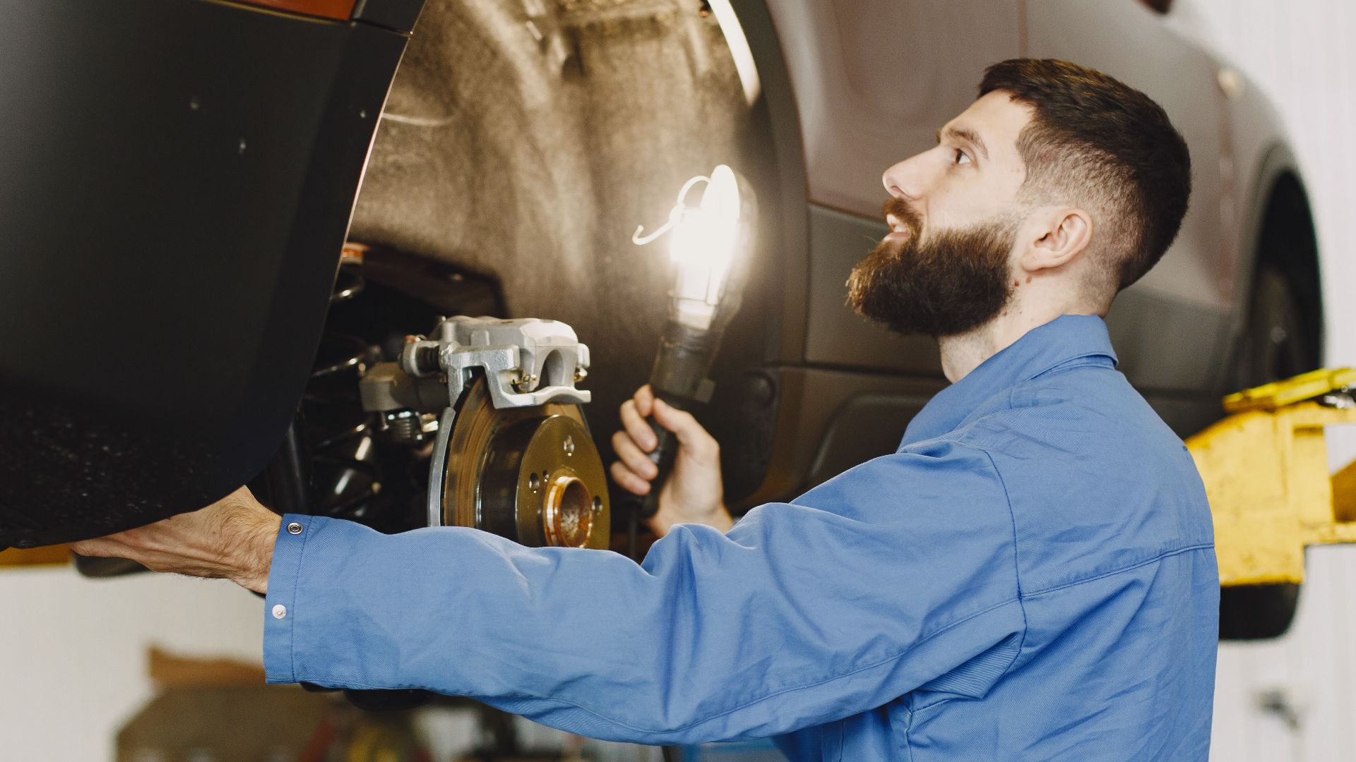 Professional mechanic in blue coveralls inspecting car brakes in a well-lit garage.