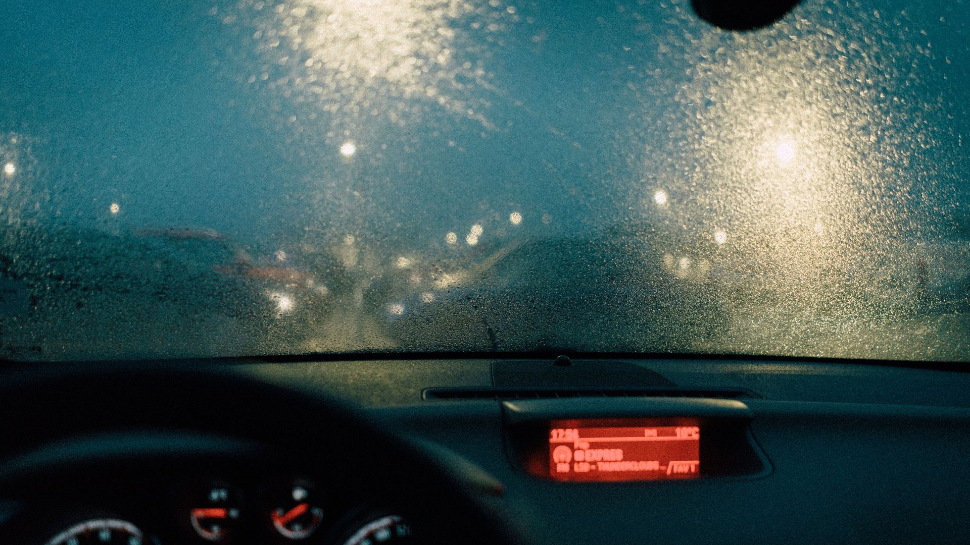 View from car interior with raindrops on windshield and blurred lights at night.