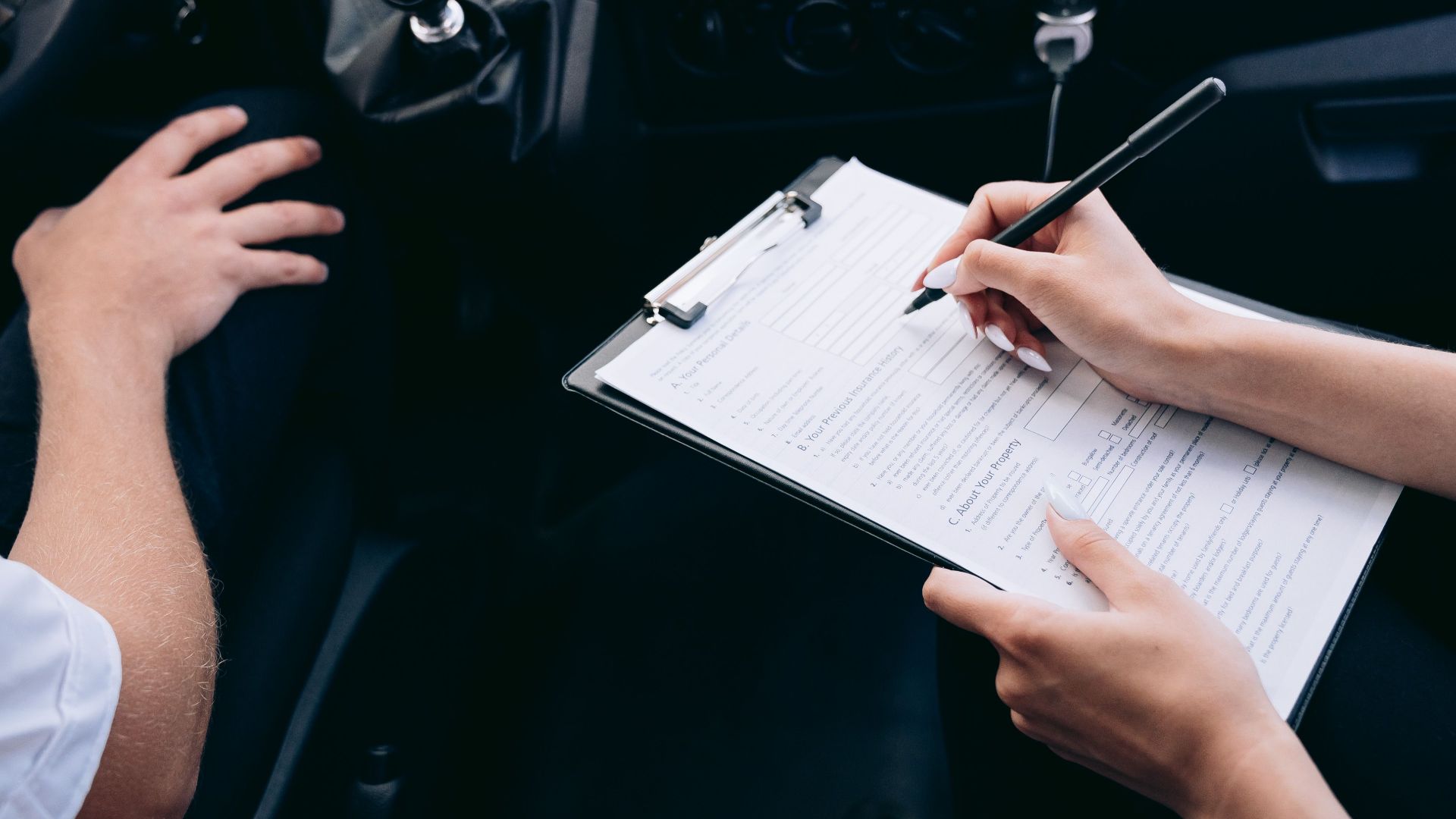 Close-up of a person writing on a clipboard inside a car, showing hands and a gear shift.