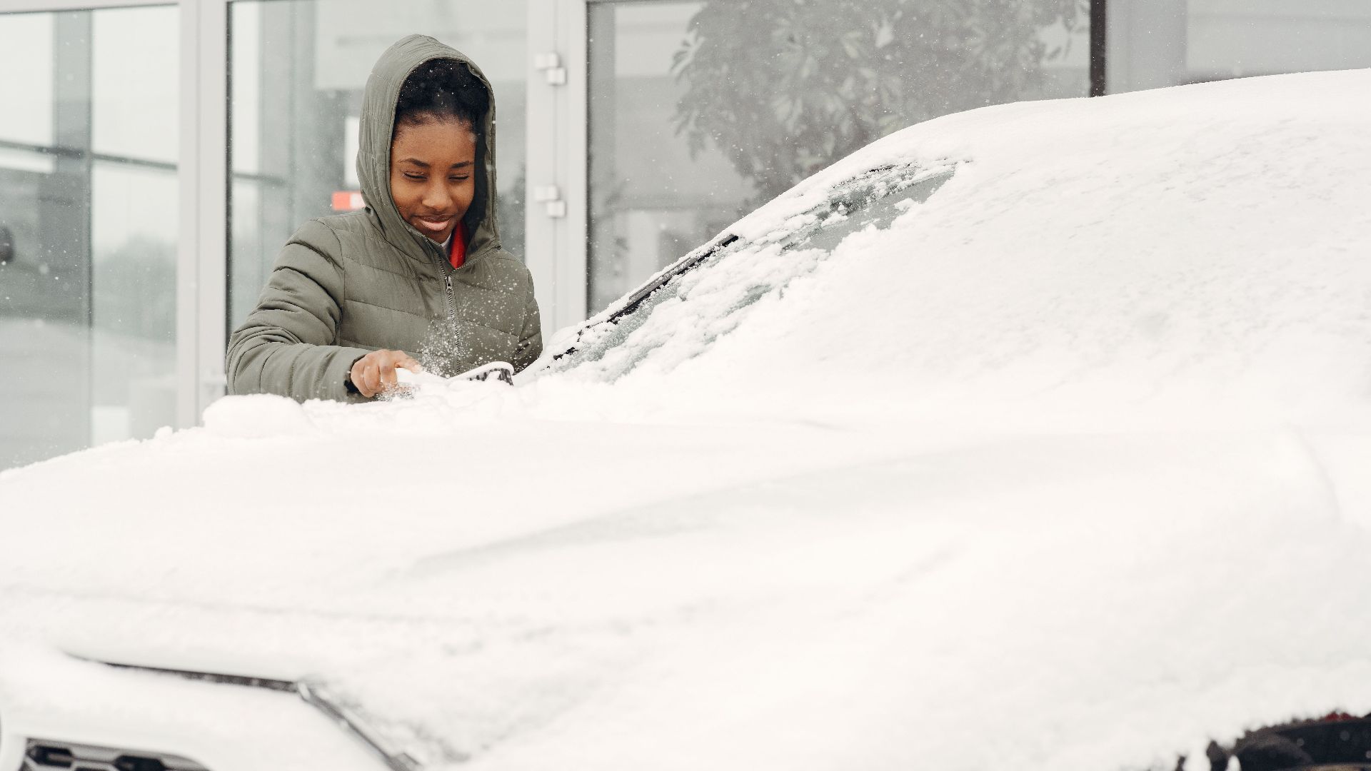 A woman in a gray coat clears snow from a car with a smile during a winter day outdoor.