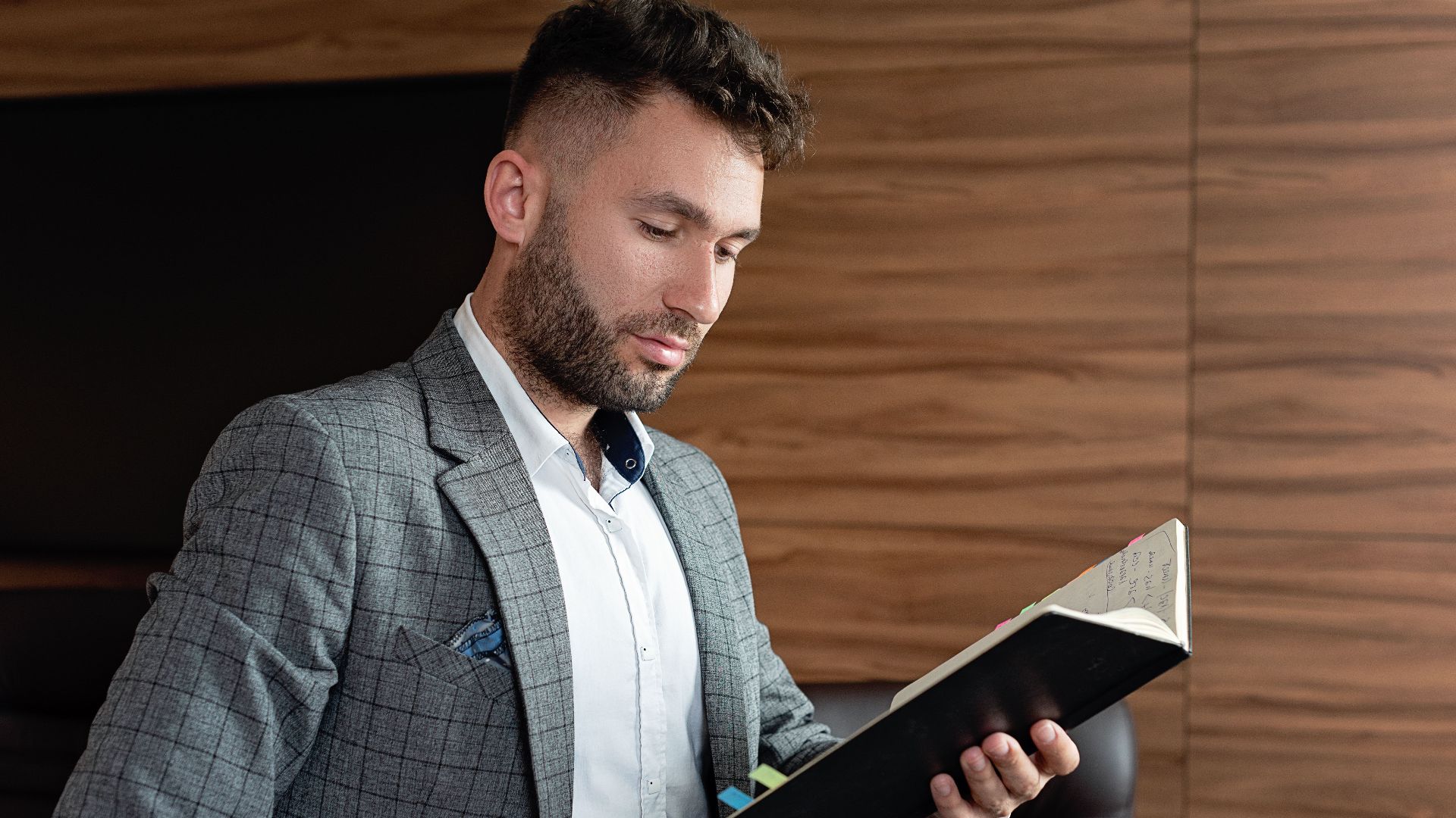 Professional businessman in office setting reading a notebook, dressed in a gray plaid blazer.