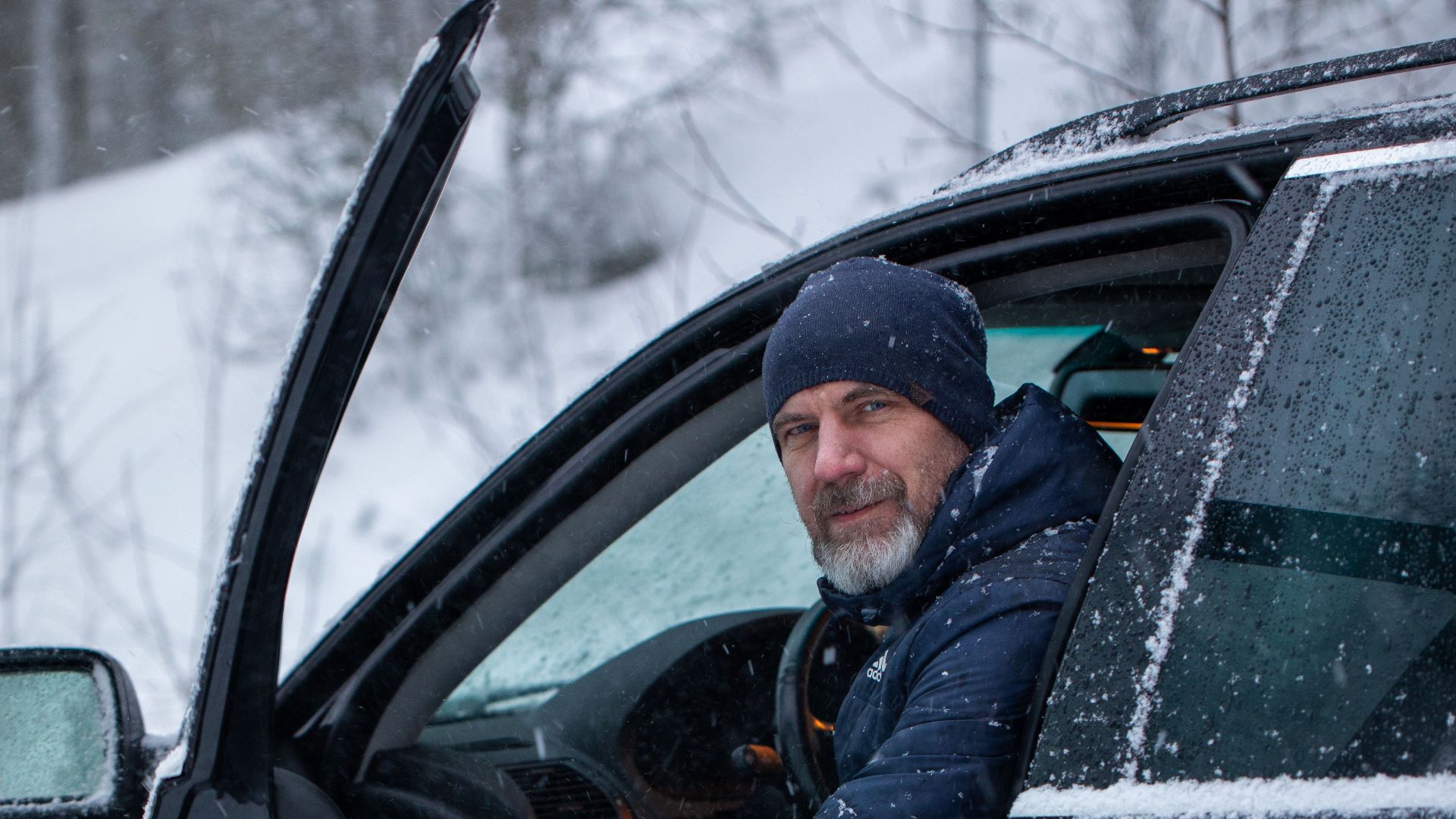 A man in winter attire sits in a car doorway amid falling snow, capturing a serene winter moment.