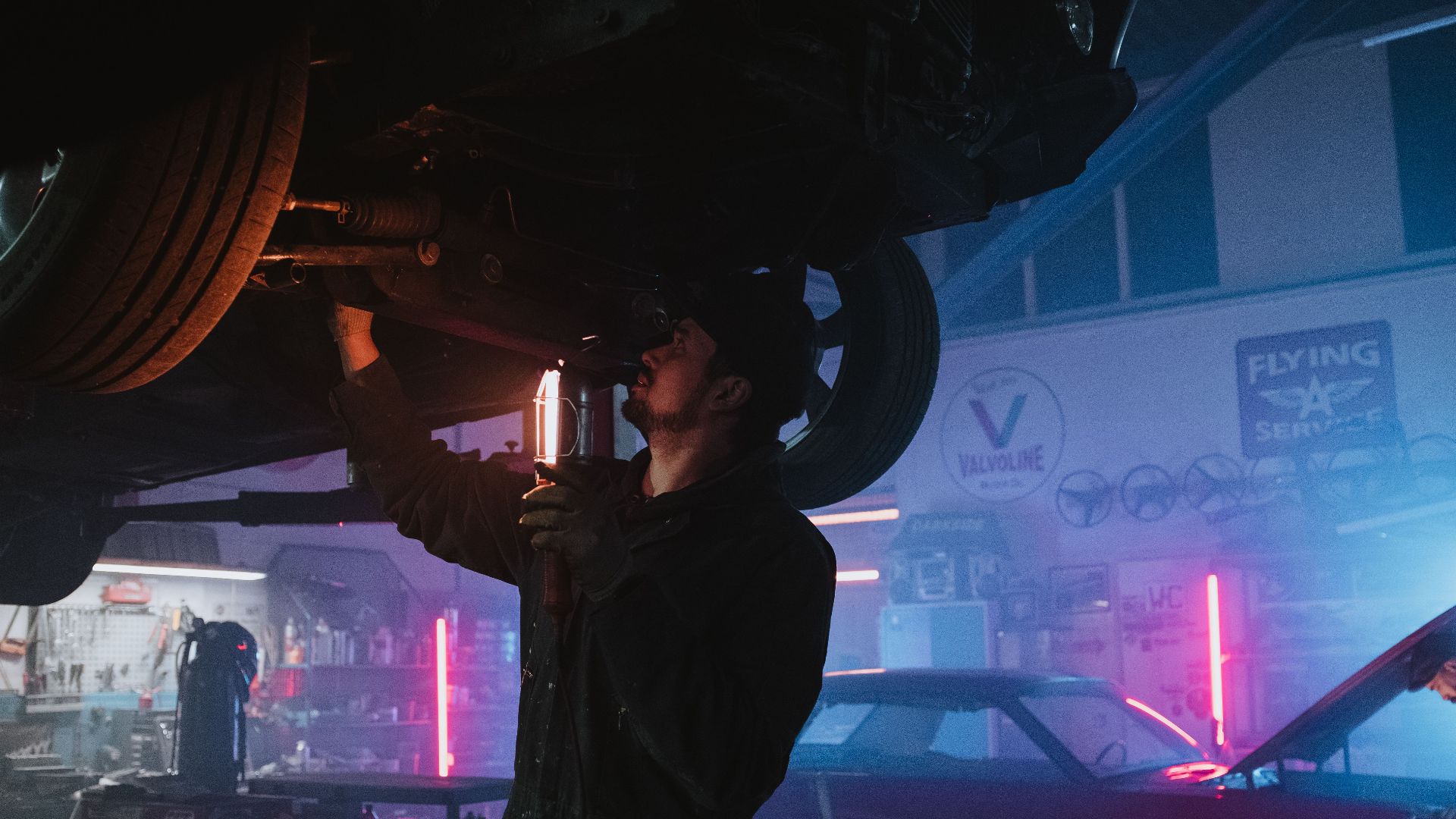 A mechanic inspects a car's undercarriage in a vibrant, neon-lit garage setting, highlighting automotive repair work.