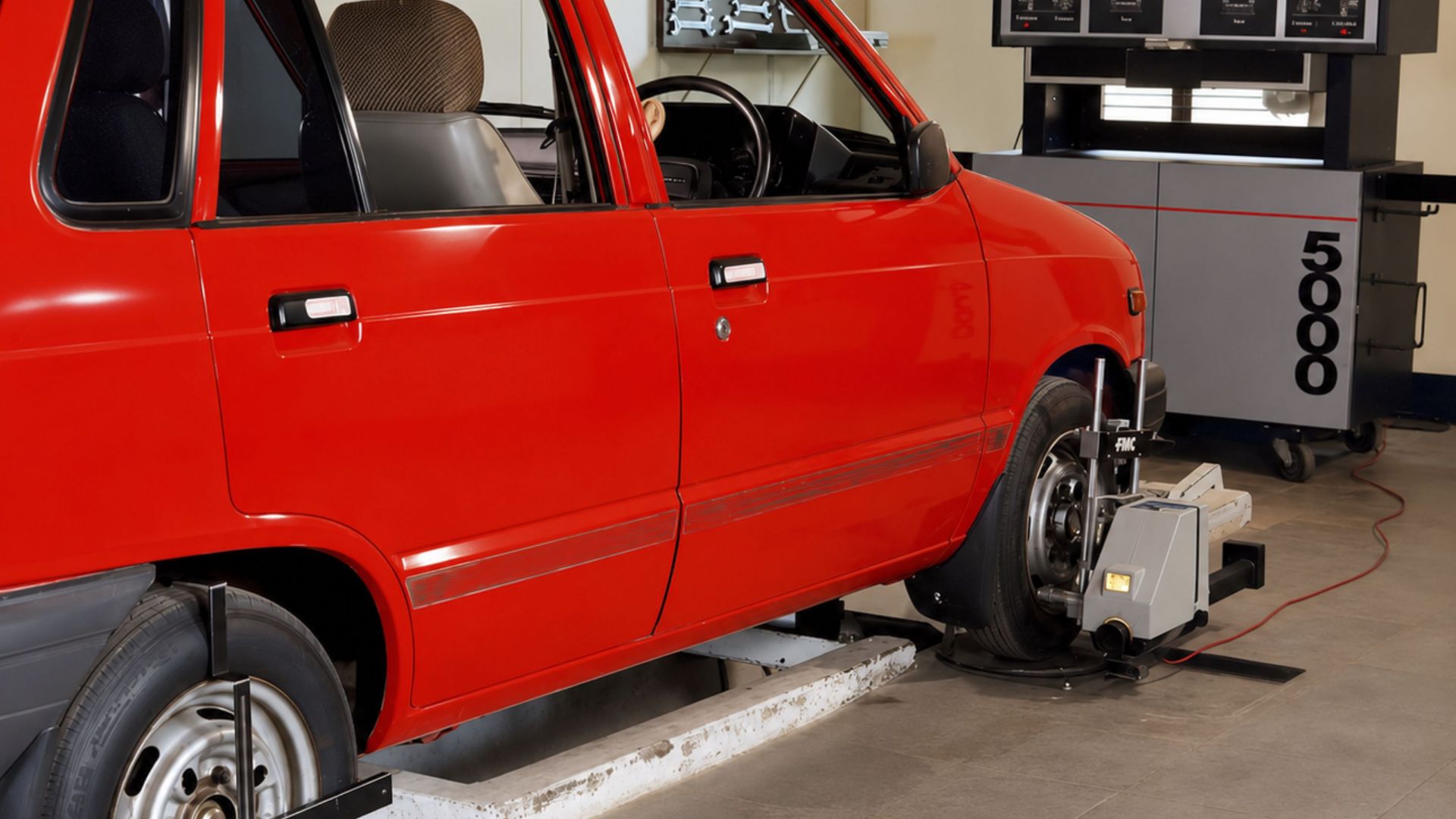 Red compact car in an automotive workshop undergoing computerized wheel alignment service.